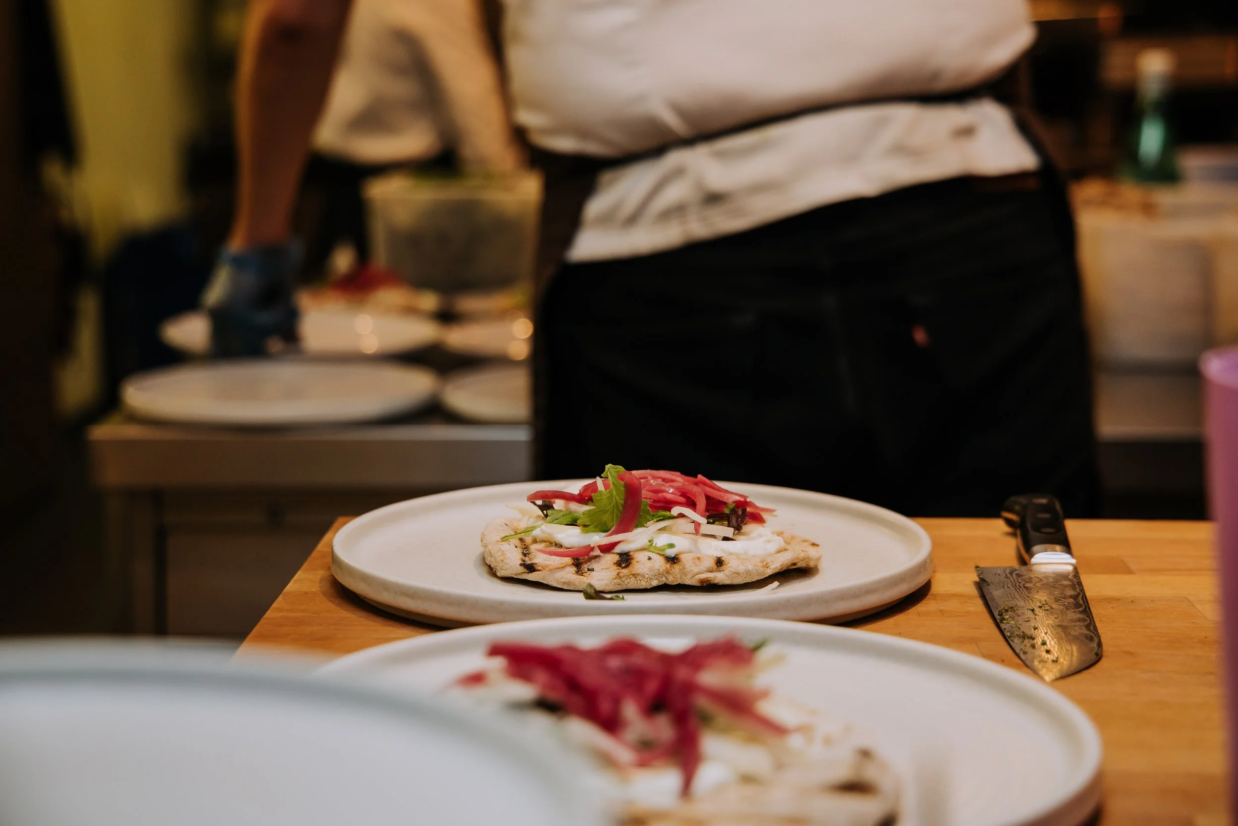 Close-up of a pizza with vegetables on a white plate, with a chef in the background in a kitchen.