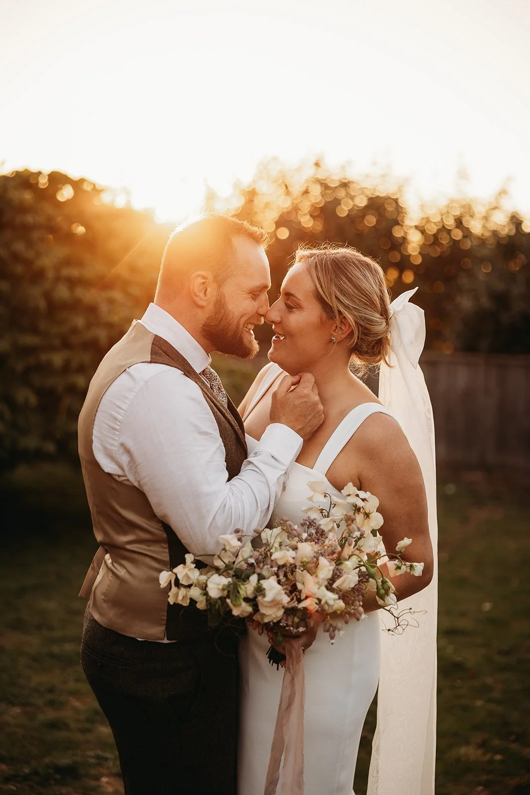 A couple on their wedding day standing close, smiling, with heads touching, outdoors at sunset. The bride is holding a bouquet of flowers and the groom is gently touching her chin.