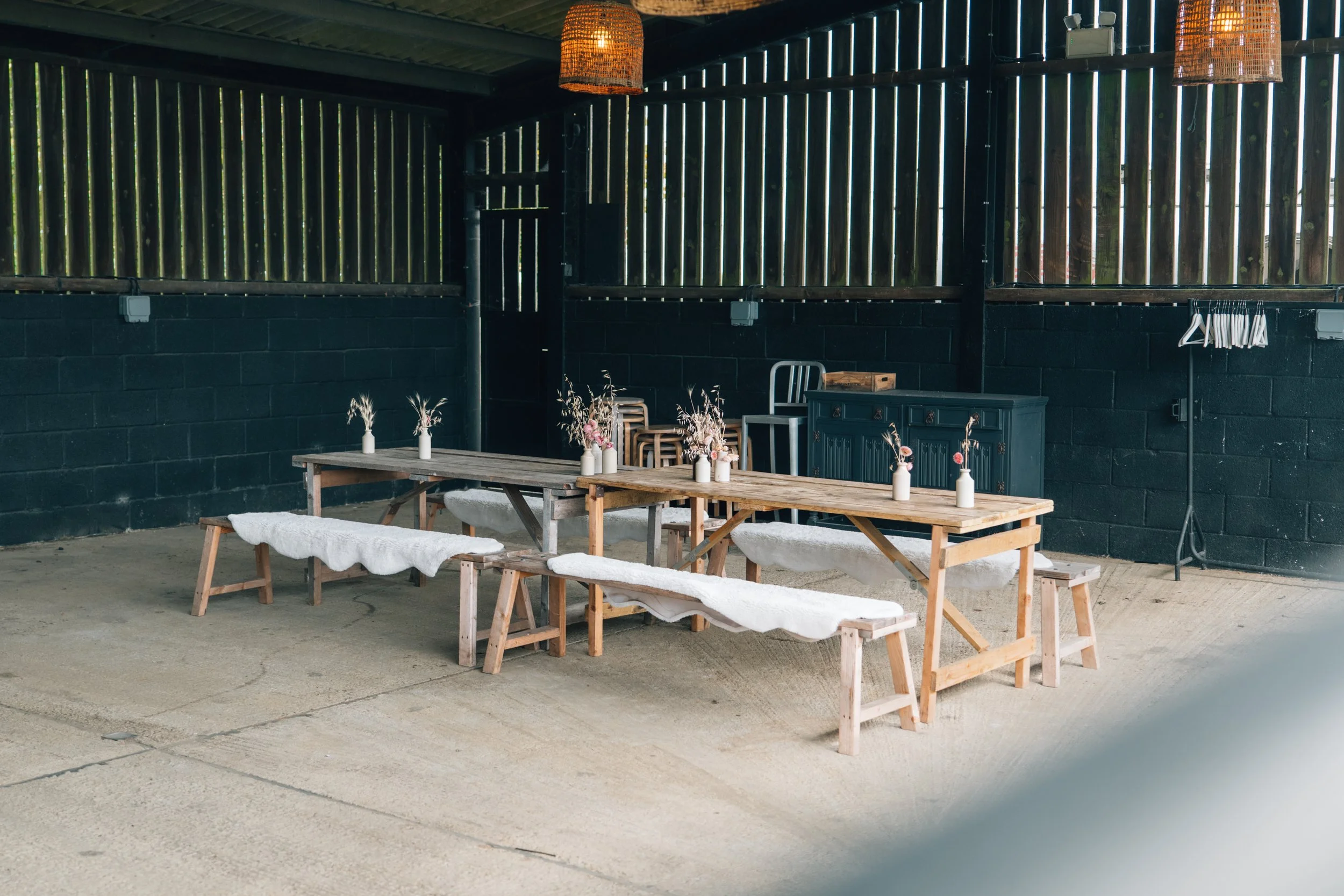 Rustic outdoor dining area with a wooden picnic table and benches covered with white fur throws, small vases with flowers, set against a black brick wall and wooden slat fence, featuring hanging pendant lights and a coat rack.