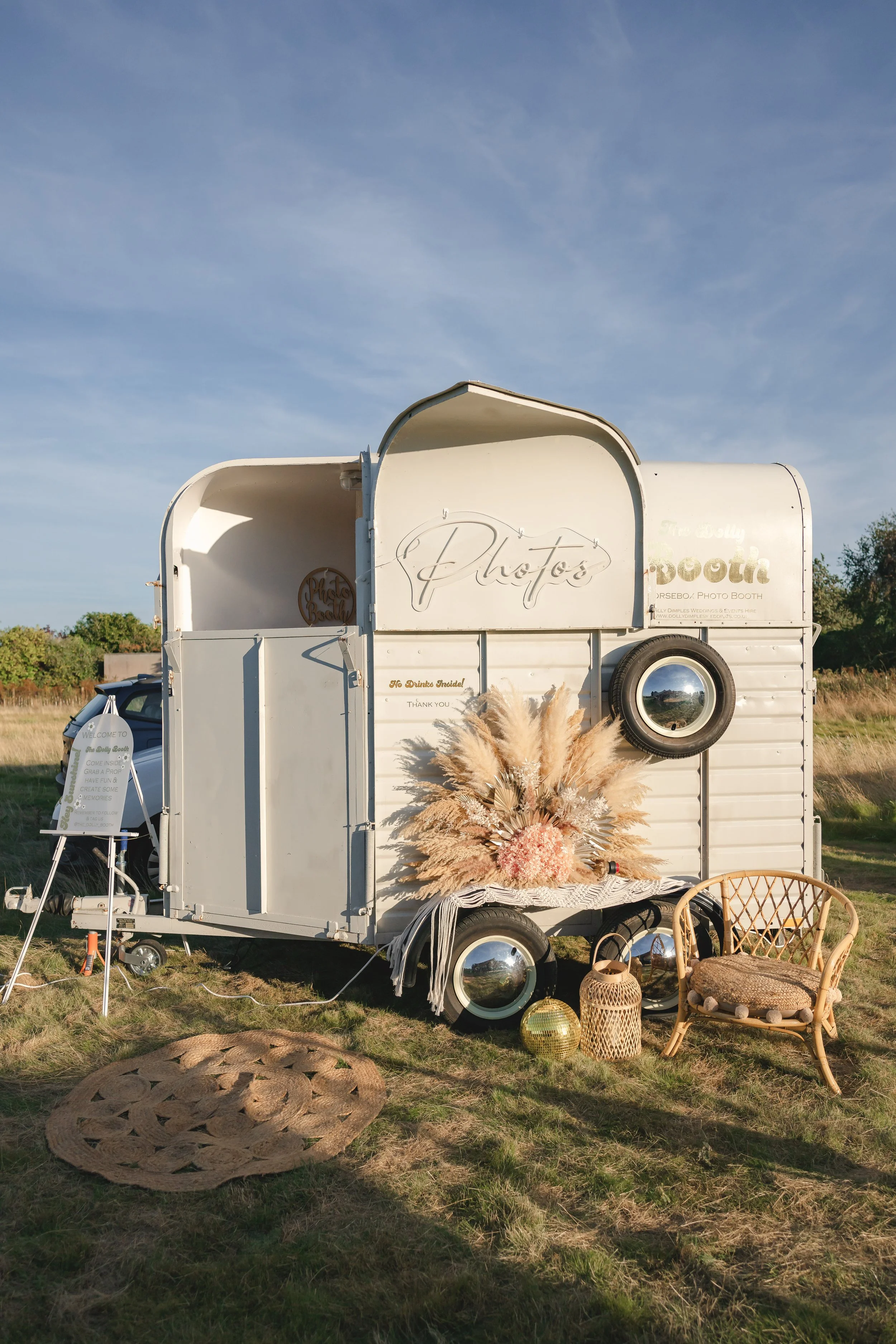 A vintage white photo booth trailer decorated with pampas grass, placed outdoors on grass with a blue sky background.
