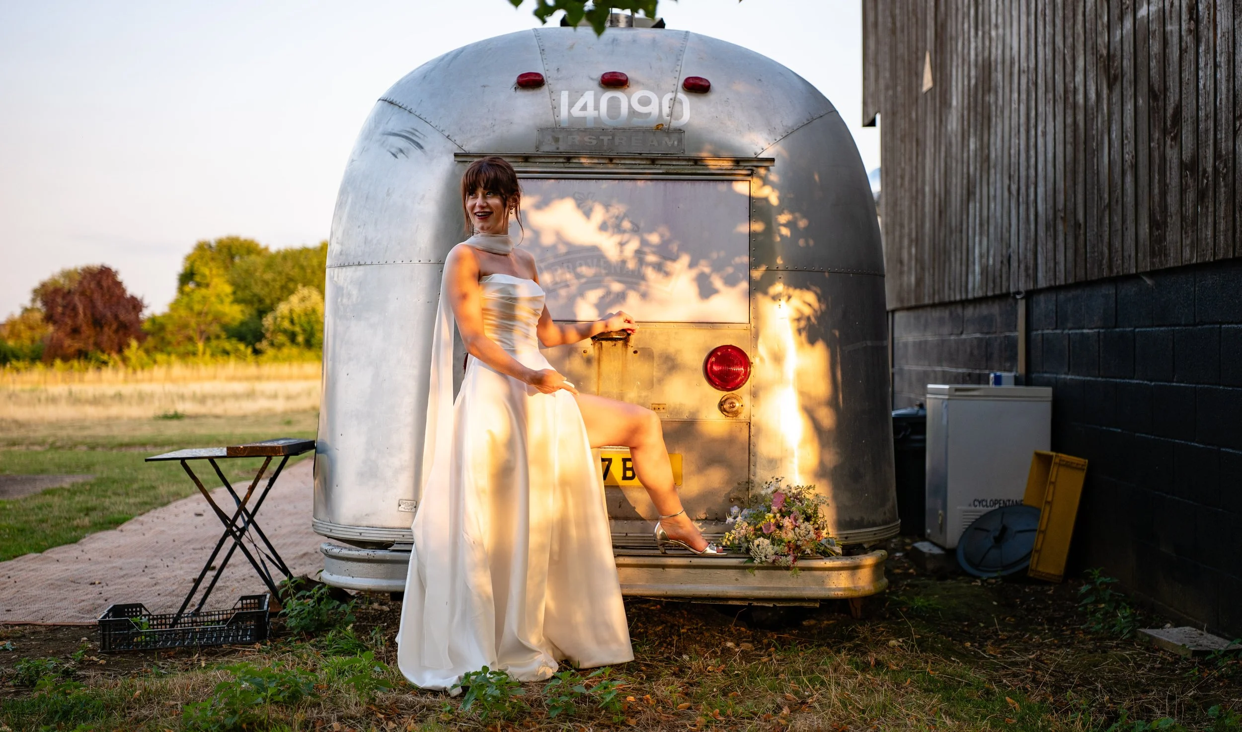 A woman in a wedding dress standing with one foot on the back of an Airstream trailer, smiling and pointing, with a bouquet on the ground nearby, outdoors during sunset.