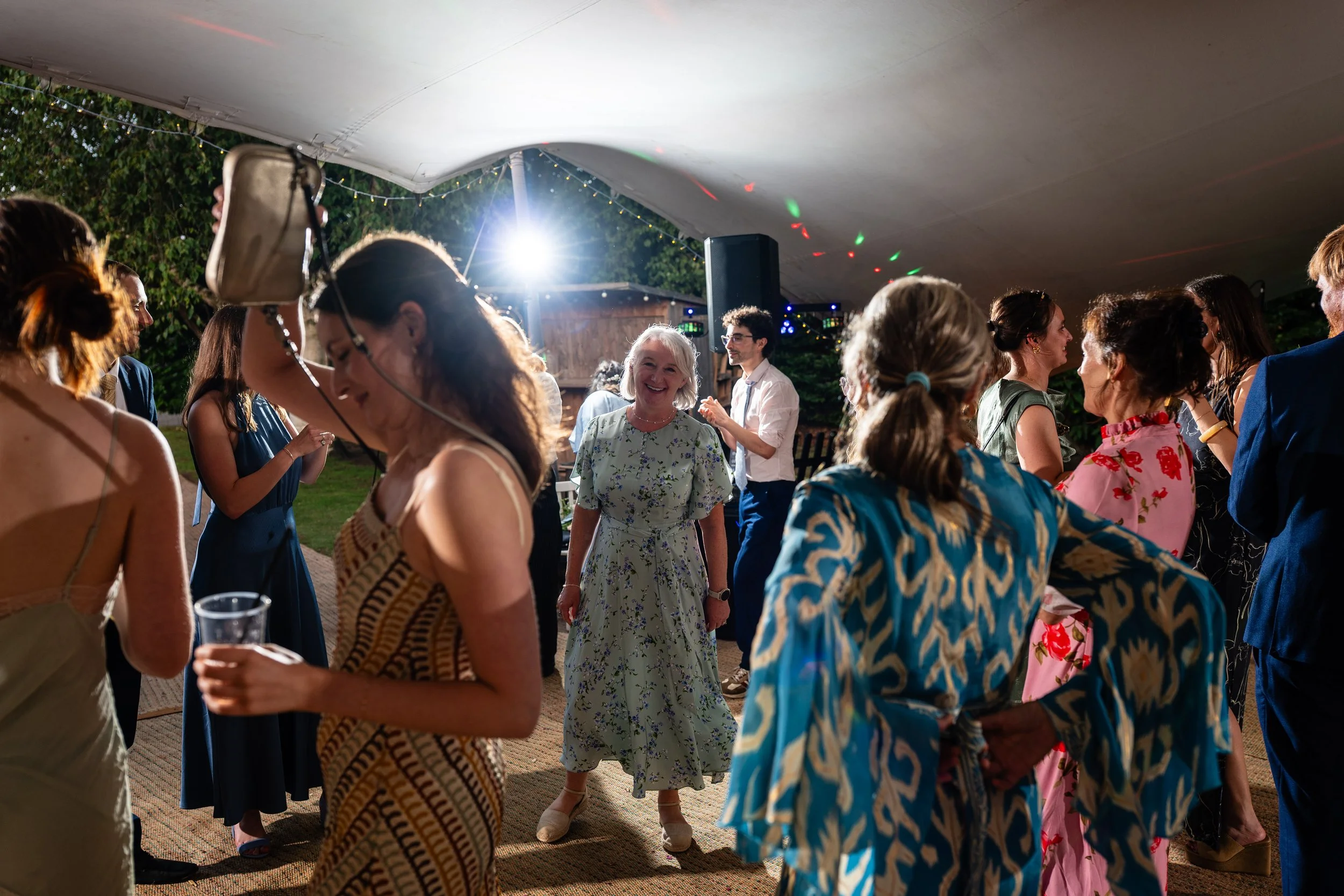 People dancing and socializing under a large tent at an outdoor event or party during nighttime with string lights and a bright light source.