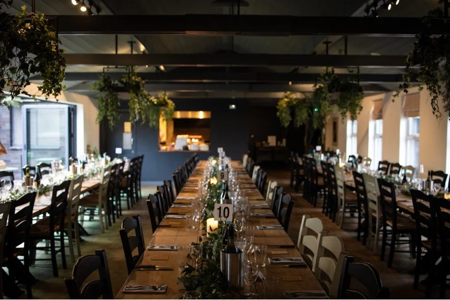 Interior of a banquet hall set up for a formal dinner with long tables, chairs, floral centerpieces, and candle lighting