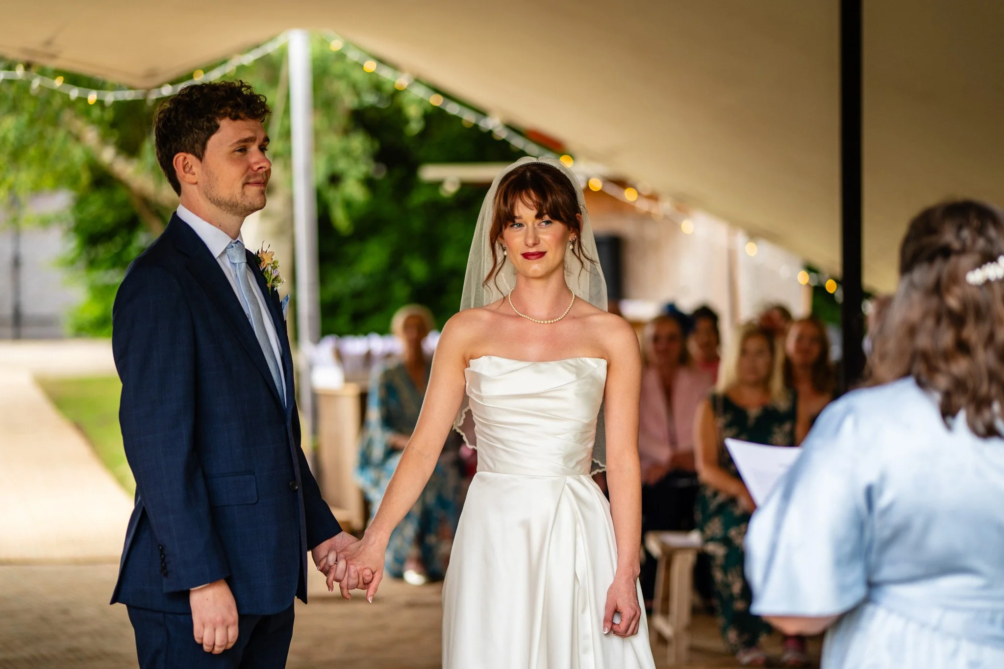 Bride and groom holding hands during wedding ceremony, with guests seated under a canopy in the background.