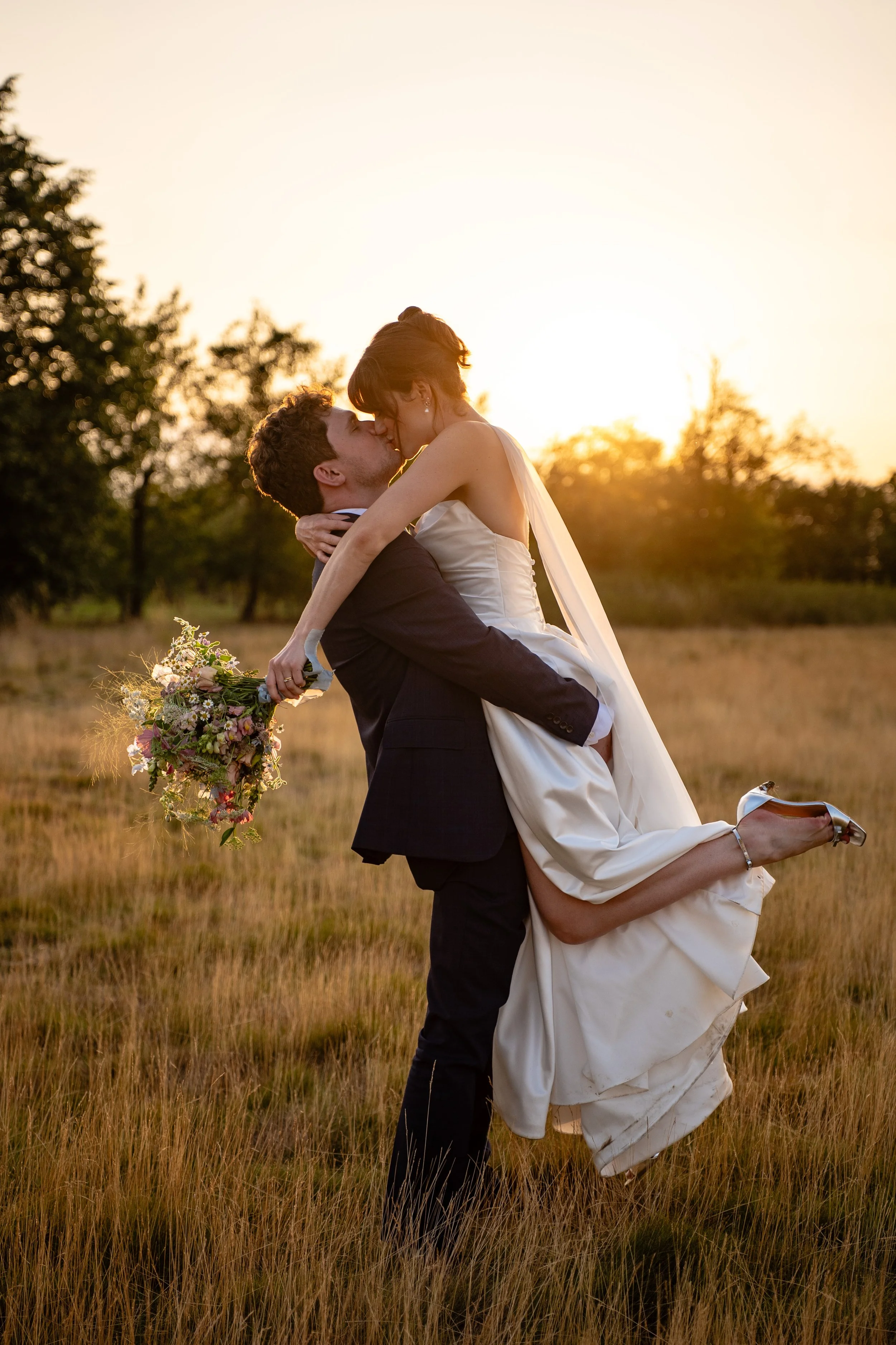 A newlywed couple in wedding attire kiss outdoors during sunset, with the groom lifting the bride, holding a bouquet of flowers.