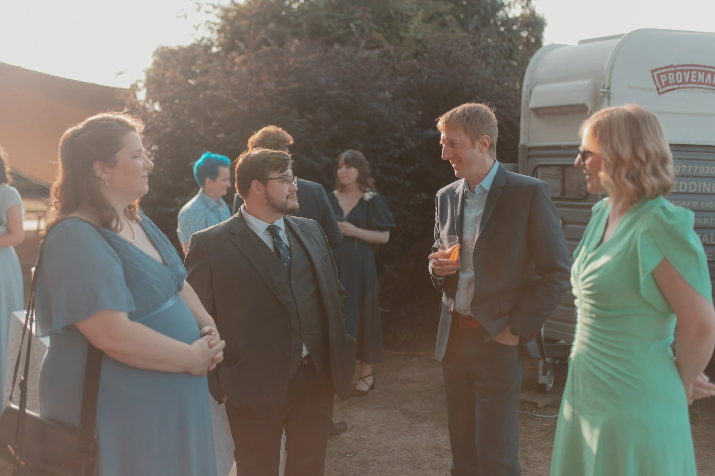A group of people dressed in semi-formal attire are outdoor during sunset, conversing and holding drinks at a social event or wedding reception, with a retro camper van in the background.