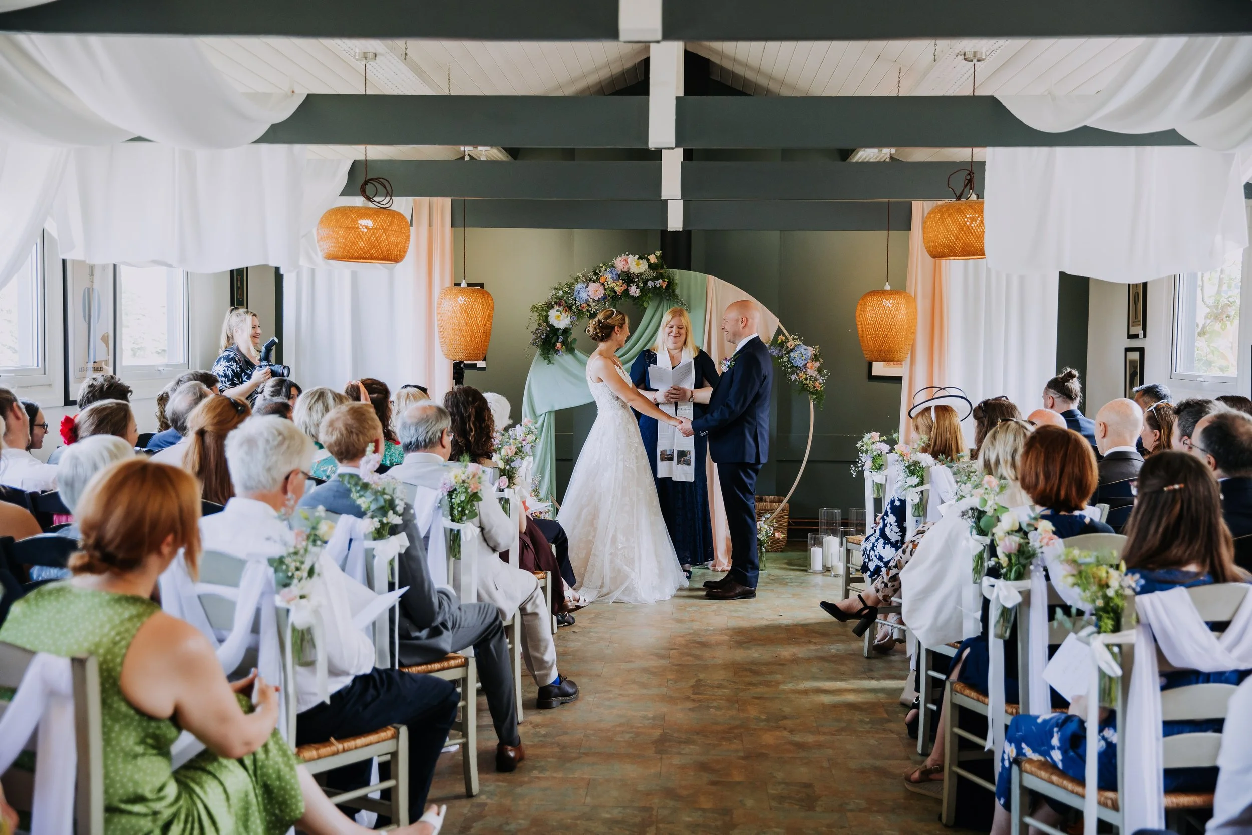 A wedding ceremony with a bride and groom facing each other, holding hands, with an officiant in the center. Guests sit on either side of the aisle, watching the couple. The setting is decorated with flowers and hanging lanterns.