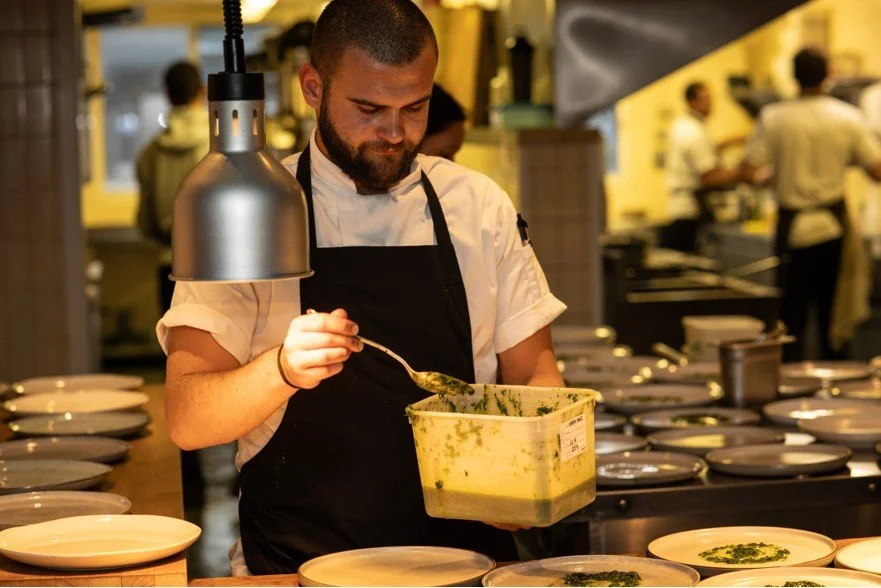 A chef in a white shirt and black apron plattering green sauce onto plates in a professional kitchen.