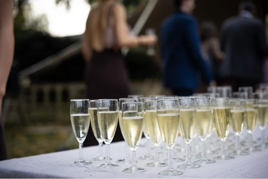 Multiple glasses of champagne on a white tablecloth at an outdoor celebration with blurred people in the background.