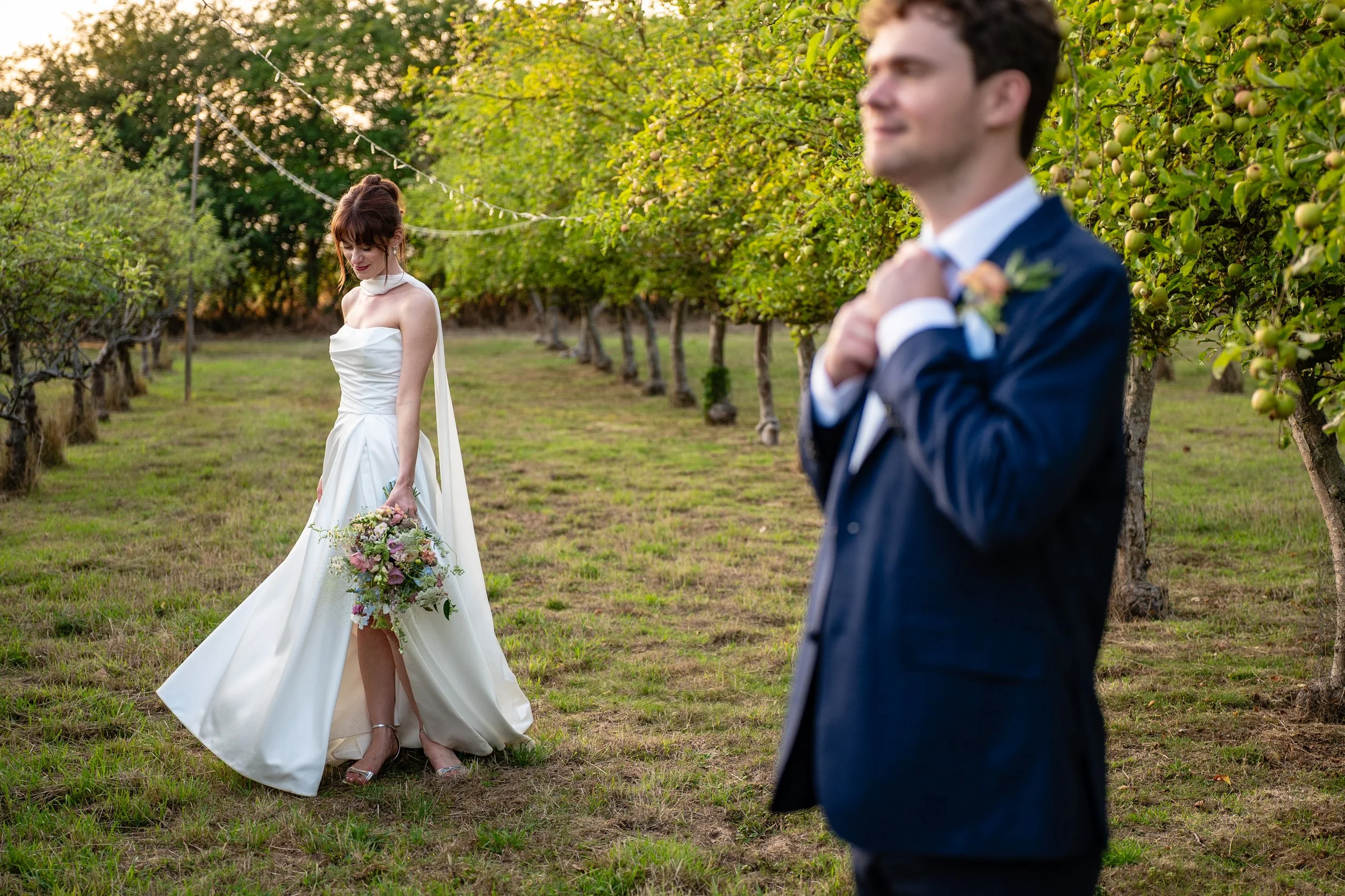 A bride in a white wedding gown holding a bouquet, standing in an orchard, looking down, while a groom in a navy suit is in the foreground adjusting his jacket, slightly out of focus.
