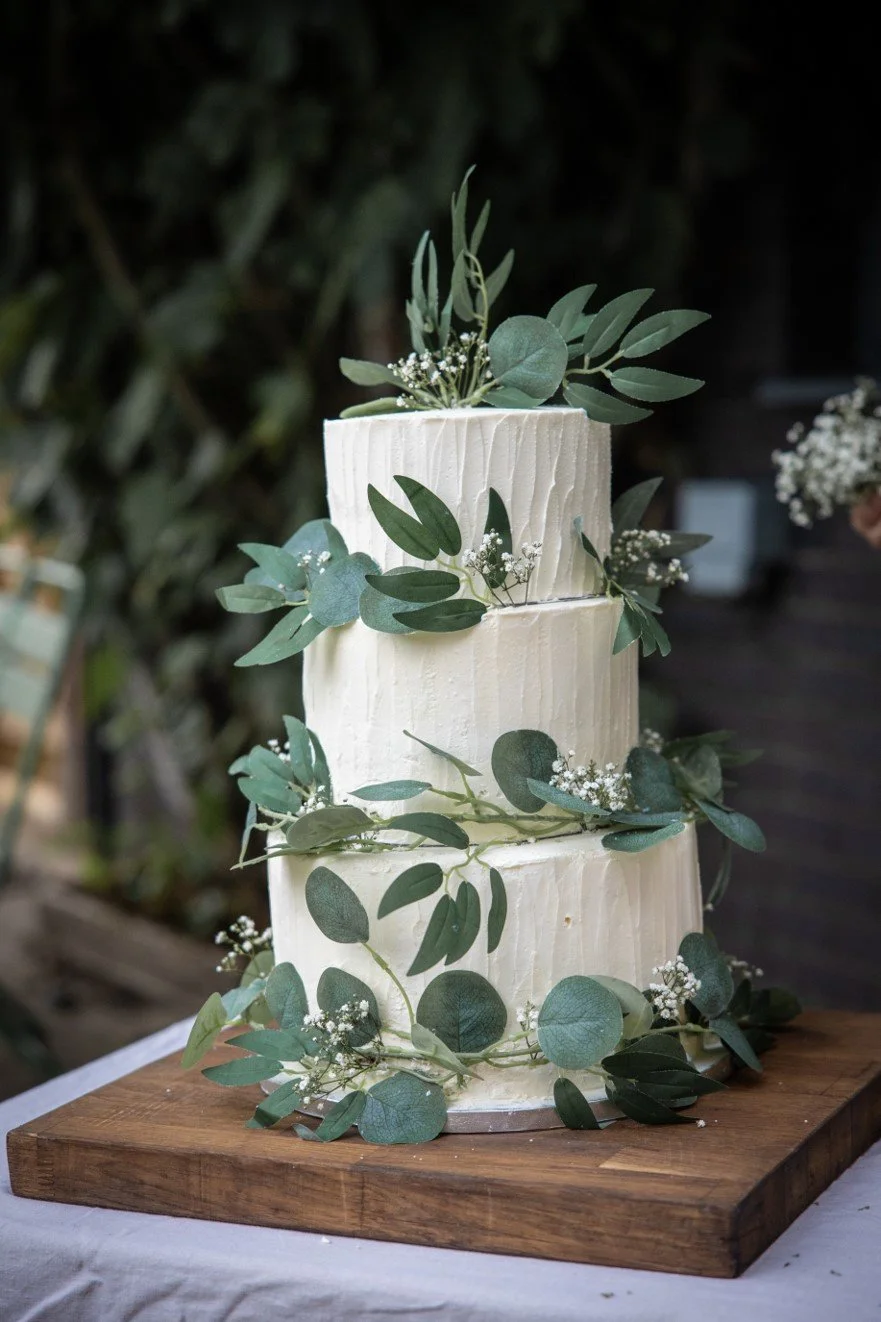 Three-tier white wedding cake decorated with green eucalyptus leaves and small white flowers, placed on a wooden stand.