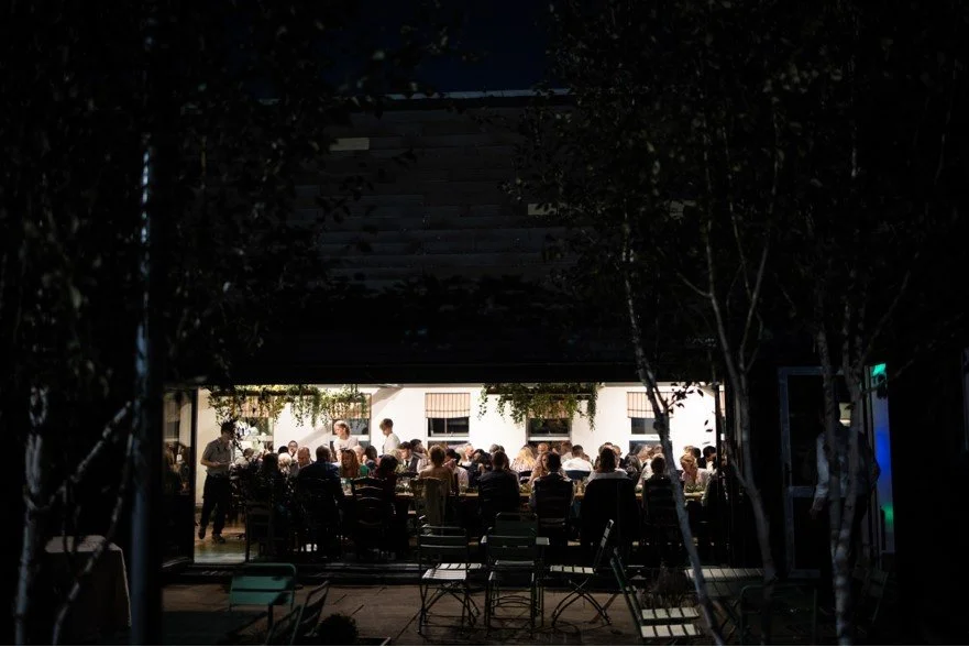 Group of people dining inside a restaurant, viewed from outside through an open window or door at night.