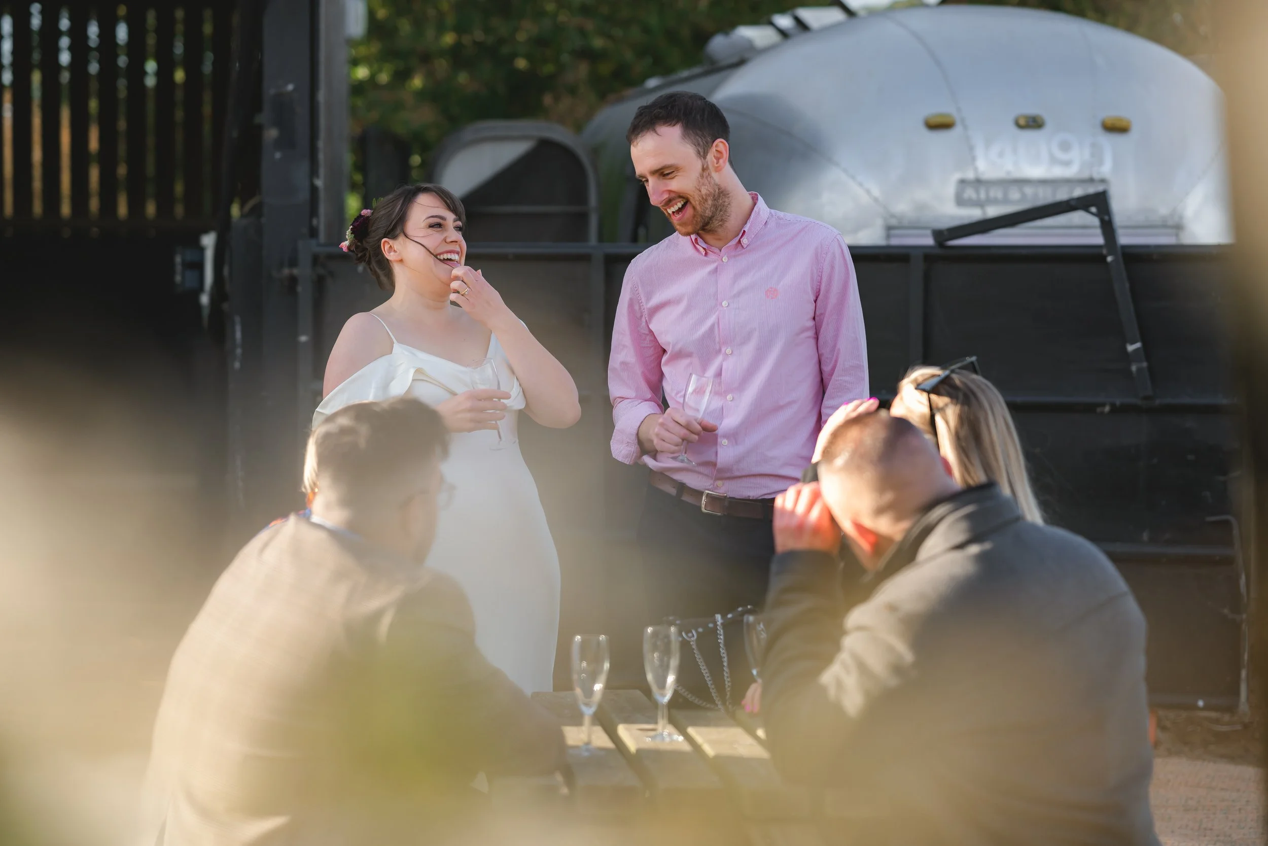 People enjoying a social outdoor gathering with drinks, smiling, and conversing near a grill or smoker in a backyard setting.