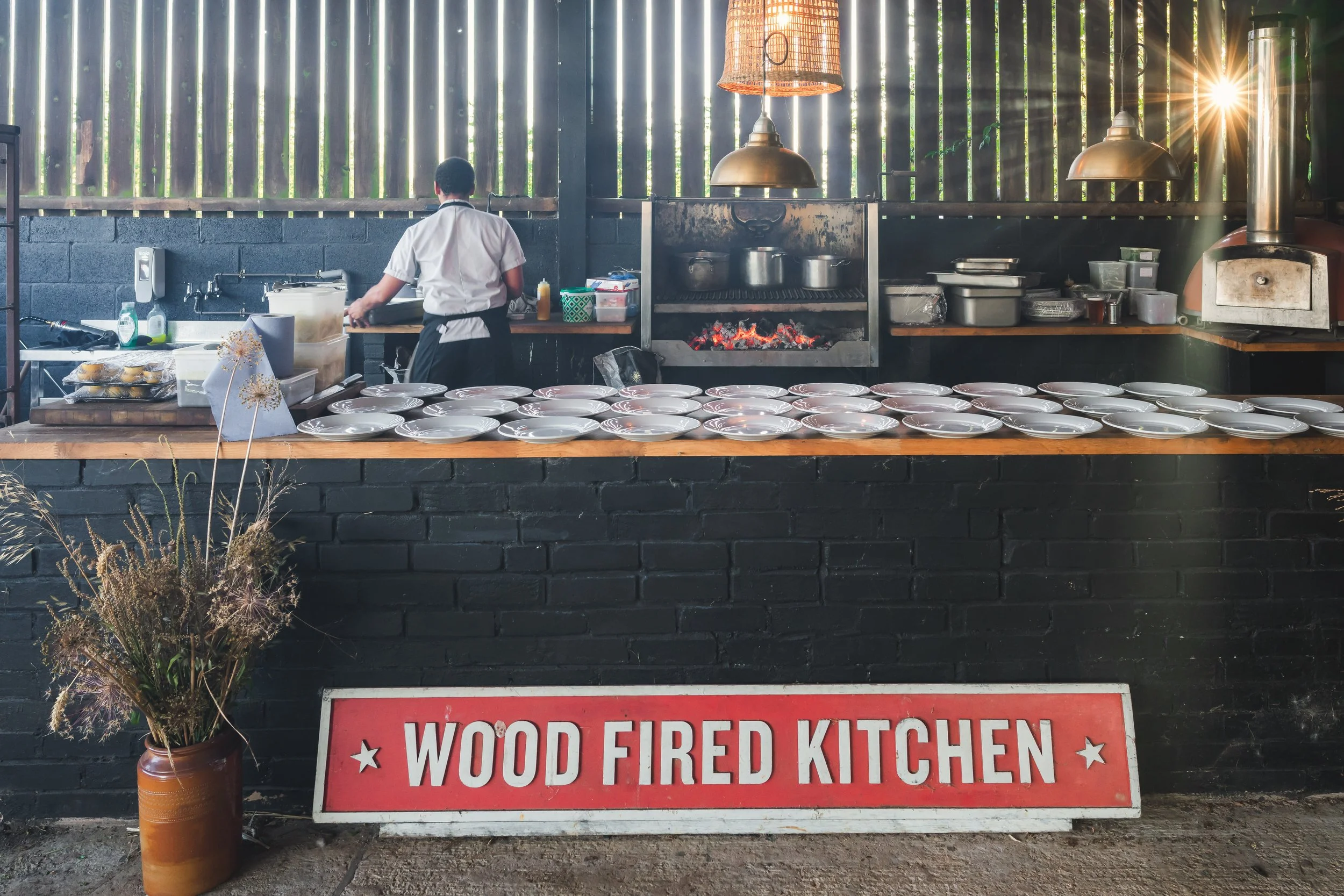 A chef working at a wood-fired kitchen, with numerous white plates arranged on a wooden counter, an open fire in the oven, and a rustic sign reading 'Wood Fired Kitchen' in front.