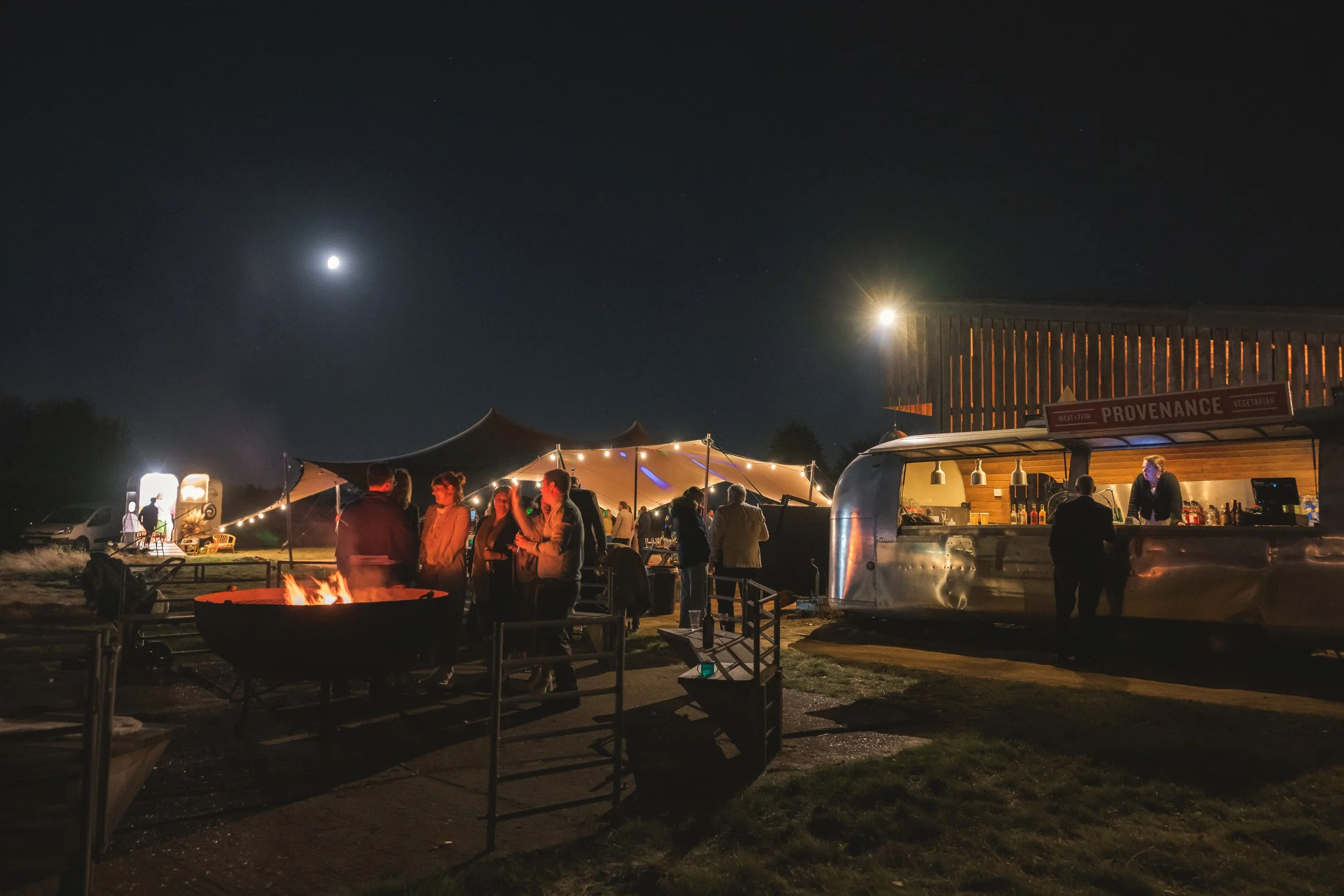 Night scene of an outdoor event with a food truck serving customers, a tent with string lights, a lit fire pit, and people socializing under a clear sky with visible moon and stars.