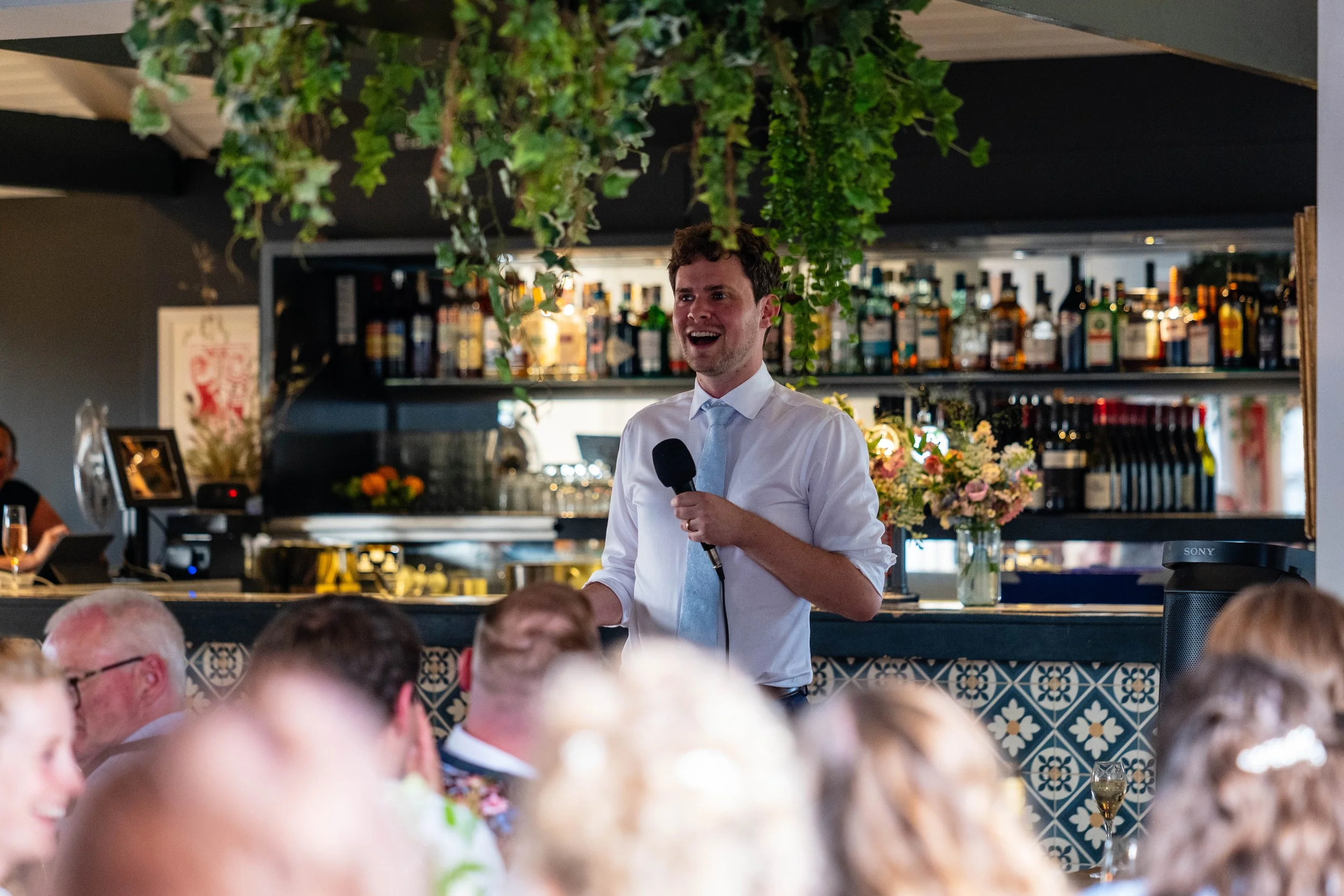 A young man in a white shirt and light gray tie giving a speech with a microphone at a social gathering, with people seated at tables and a bar with liquor bottles and flowers in the background.