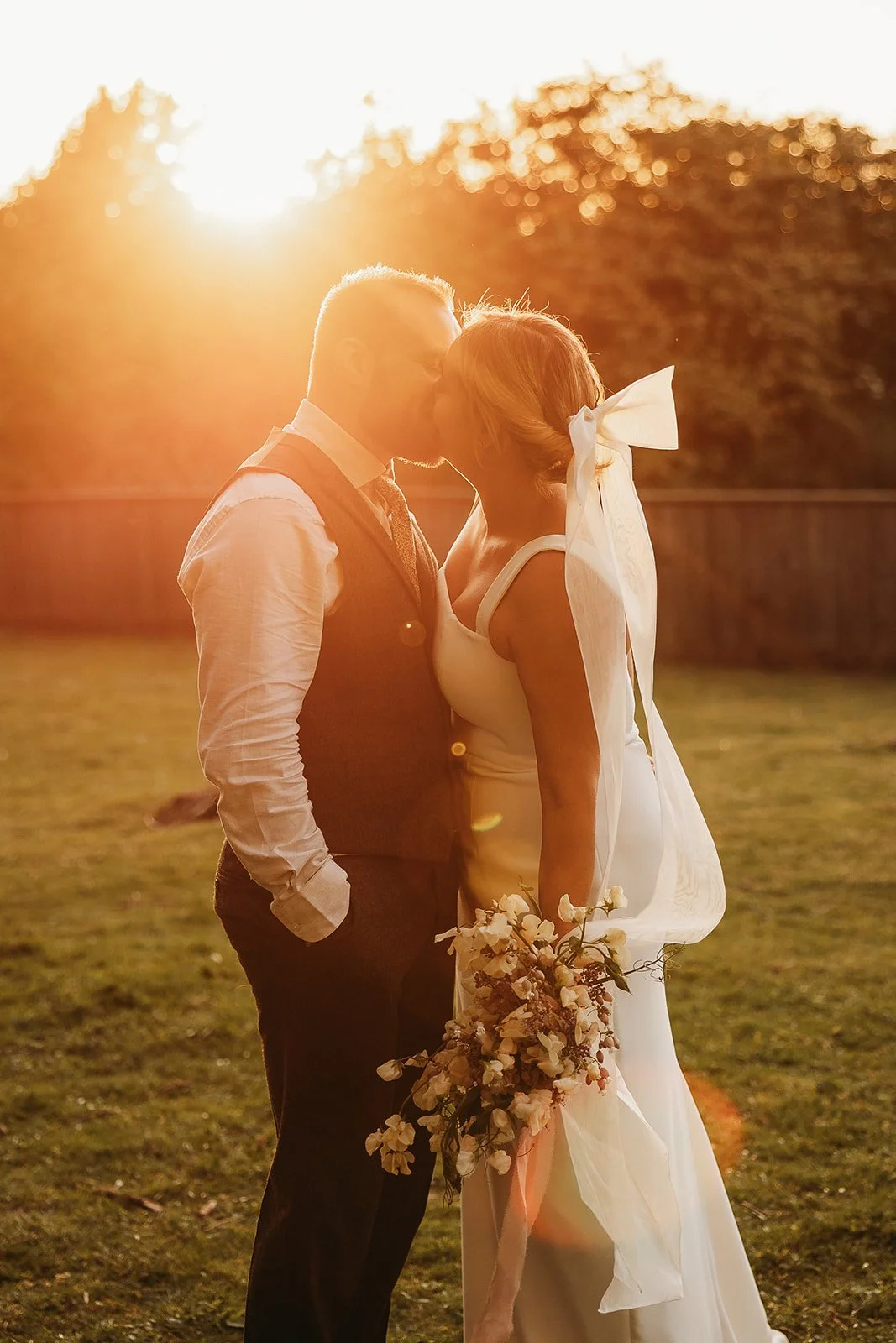A bride and groom kissing outdoors at sunset, with the bride holding a bouquet and wearing a white dress with a large bow in her hair.
