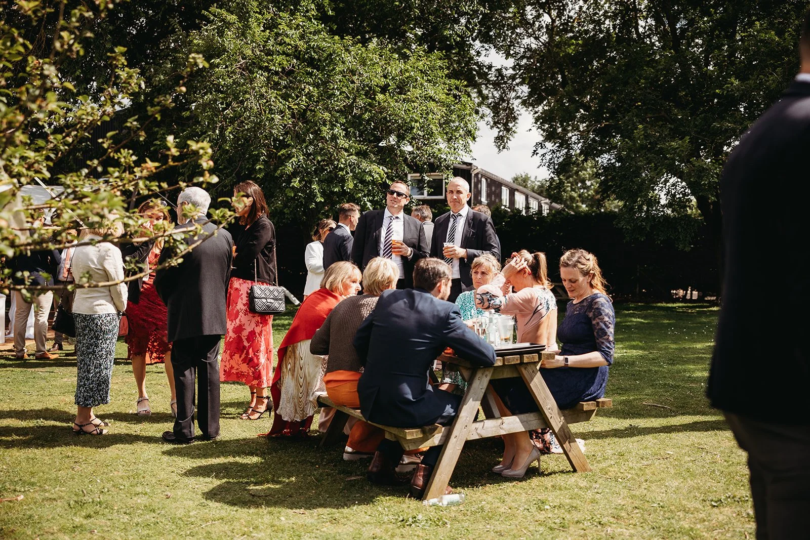People gathered outdoors at a social event, some seated at a picnic table and others standing, under trees on a sunny day.