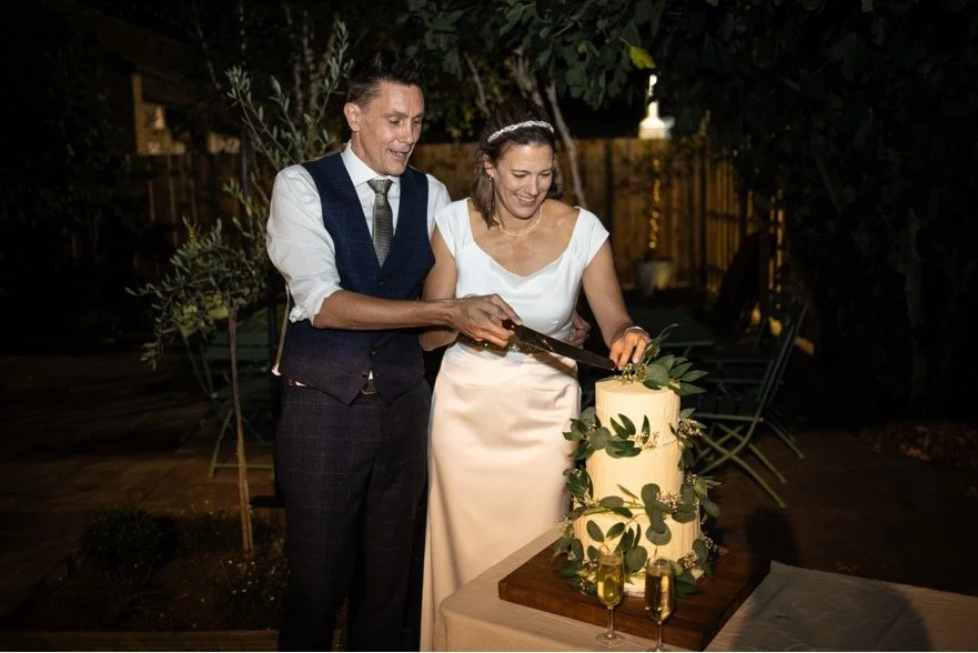 A couple in wedding attire cutting a wedding cake outdoors at night, decorated with greenery and topped with leaves, with two glasses of champagne on the table.