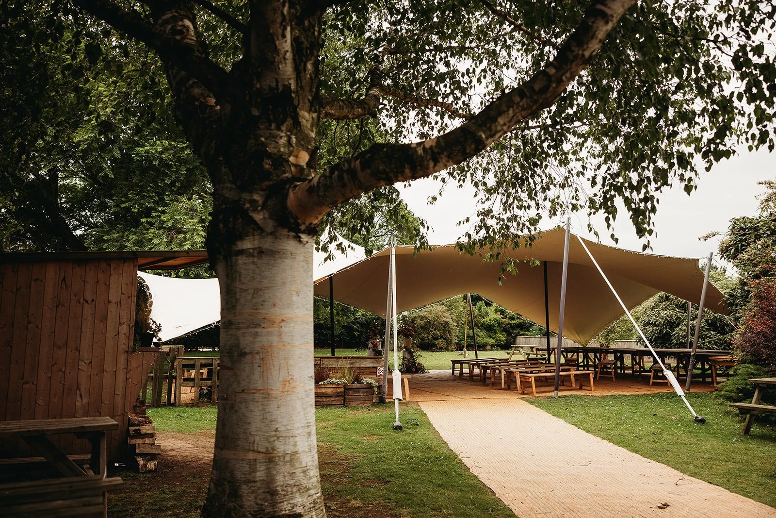 A large tree in the foreground with a wooden building to the left and an outdoor seating area with picnic tables under a beige canopy in the background.