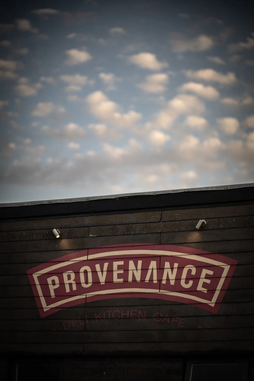 Outdoor sign for Provenance Deli Kitchen Cafe with a cloudy sky in the background.
