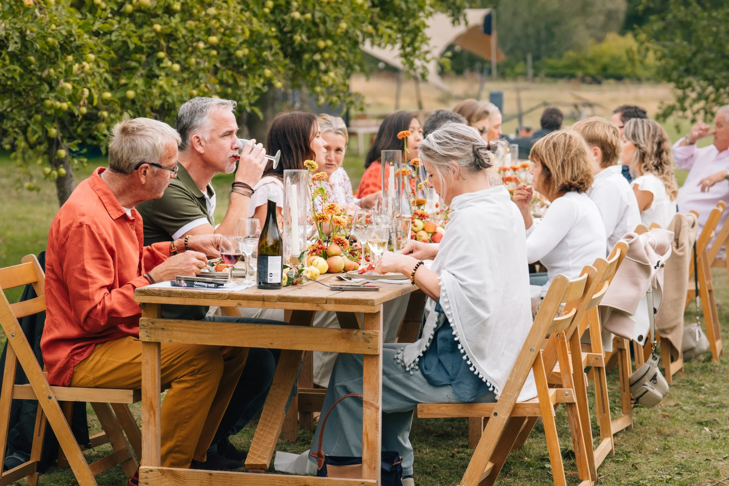 A large outdoor dinner party with people sitting at a long wooden table, drinking wine, and engaging in conversation amid a garden setting with trees and greenery.
