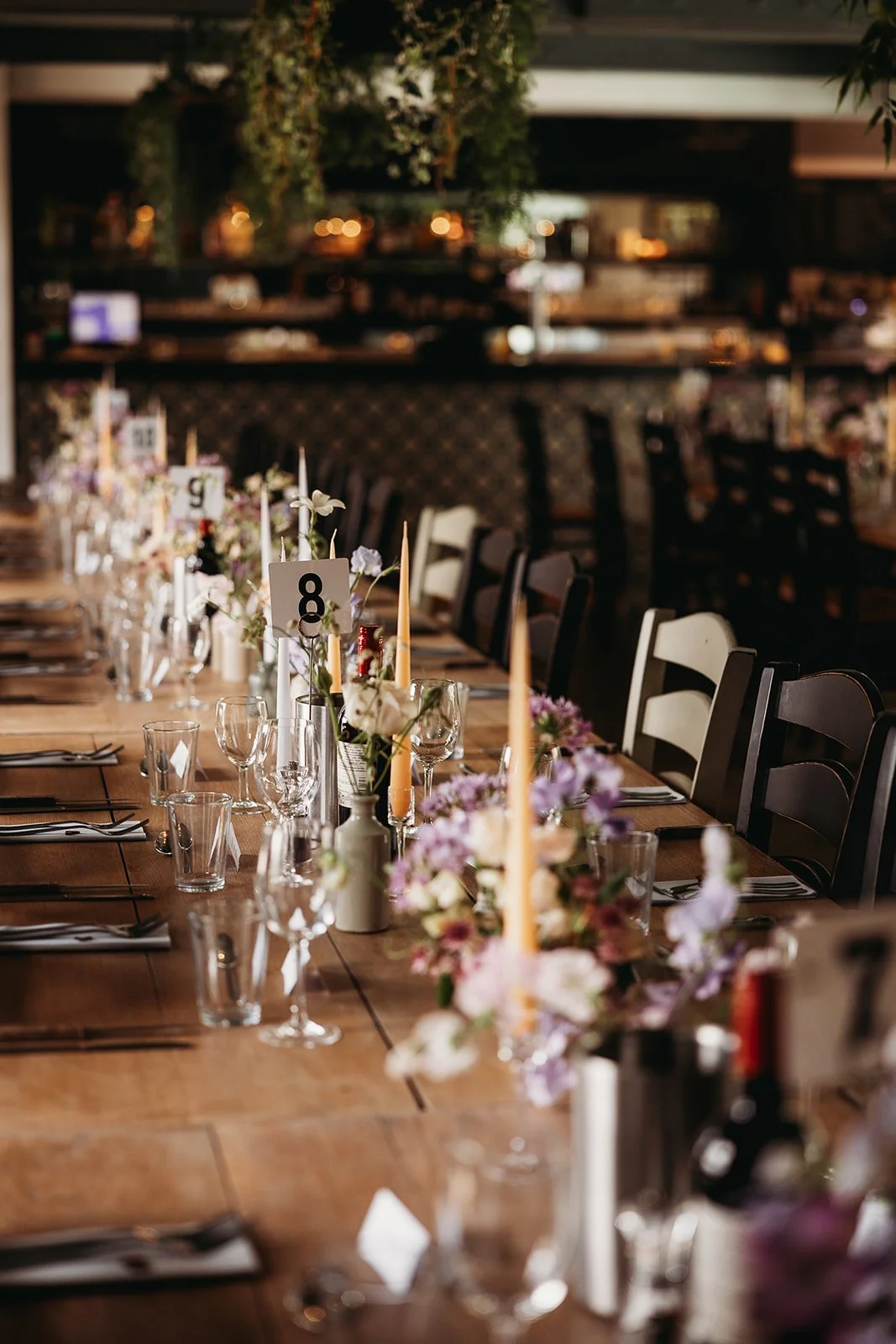 A long wooden dining table set for a formal event, decorated with flowers, candles, and glassware, with chairs arranged along both sides.
