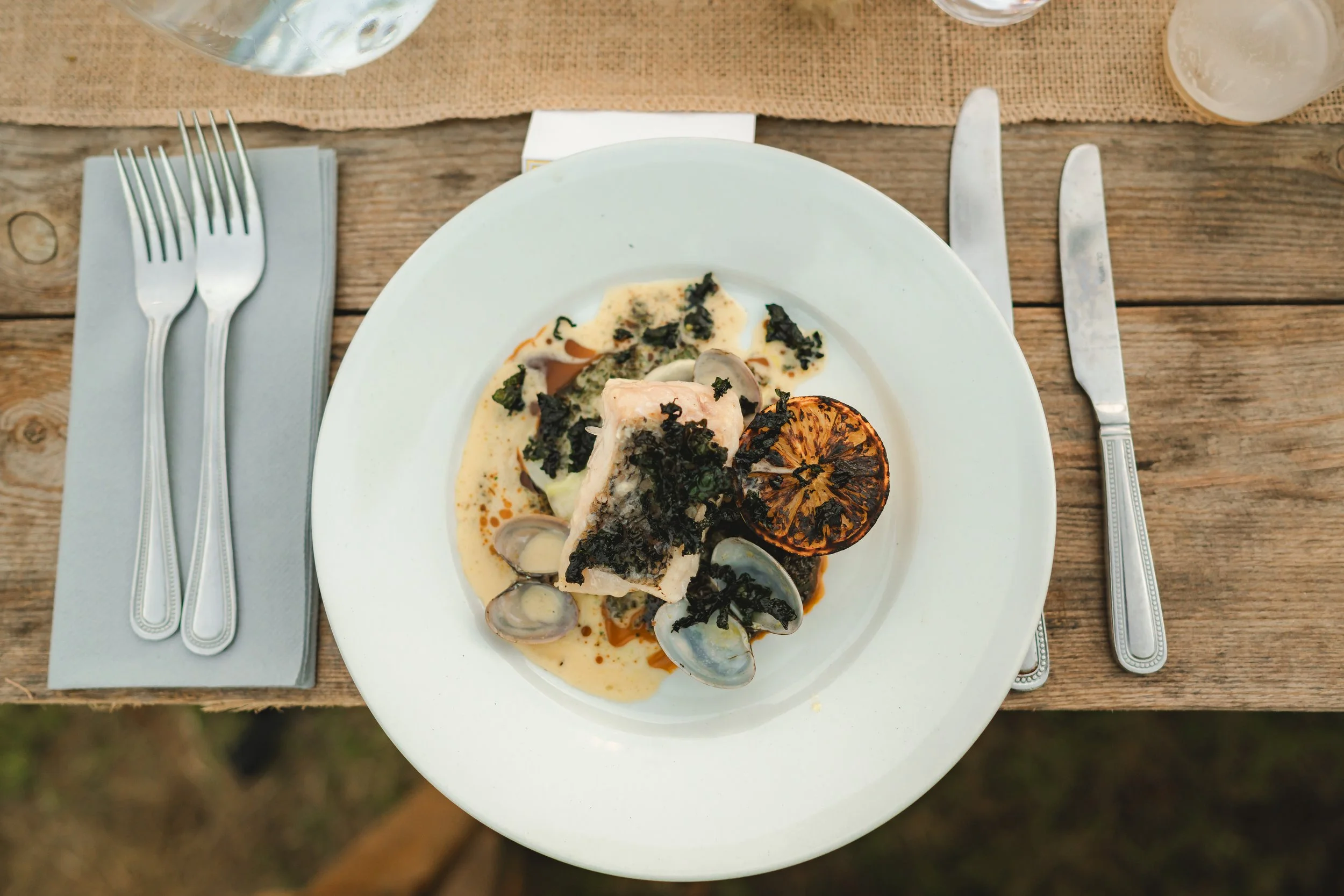 A plate of cooked fish garnished with herbs, served with clams, grilled lemon half, and sauce. The table has silverware, a napkin, and glasses of water.