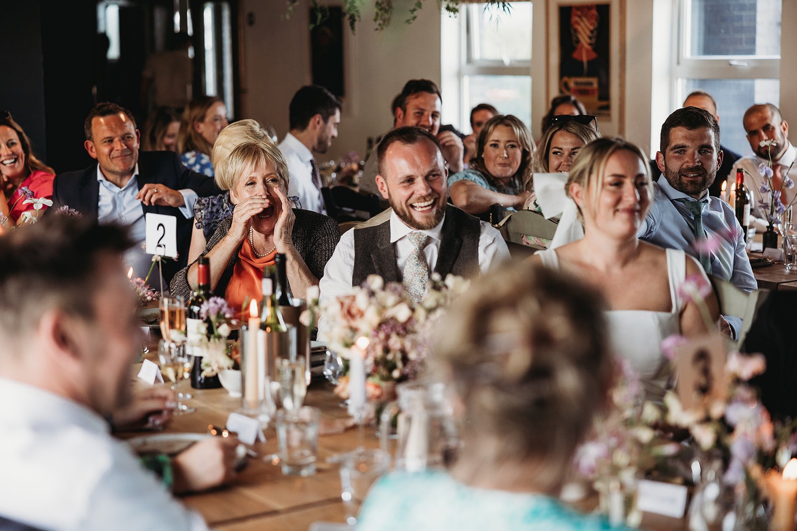 A group of people at a wedding reception, sitting at tables with floral centerpieces and decorated with candles. They are smiling and laughing, dressed in formal attire, with some raising their hands or covering their mouths in laughter.