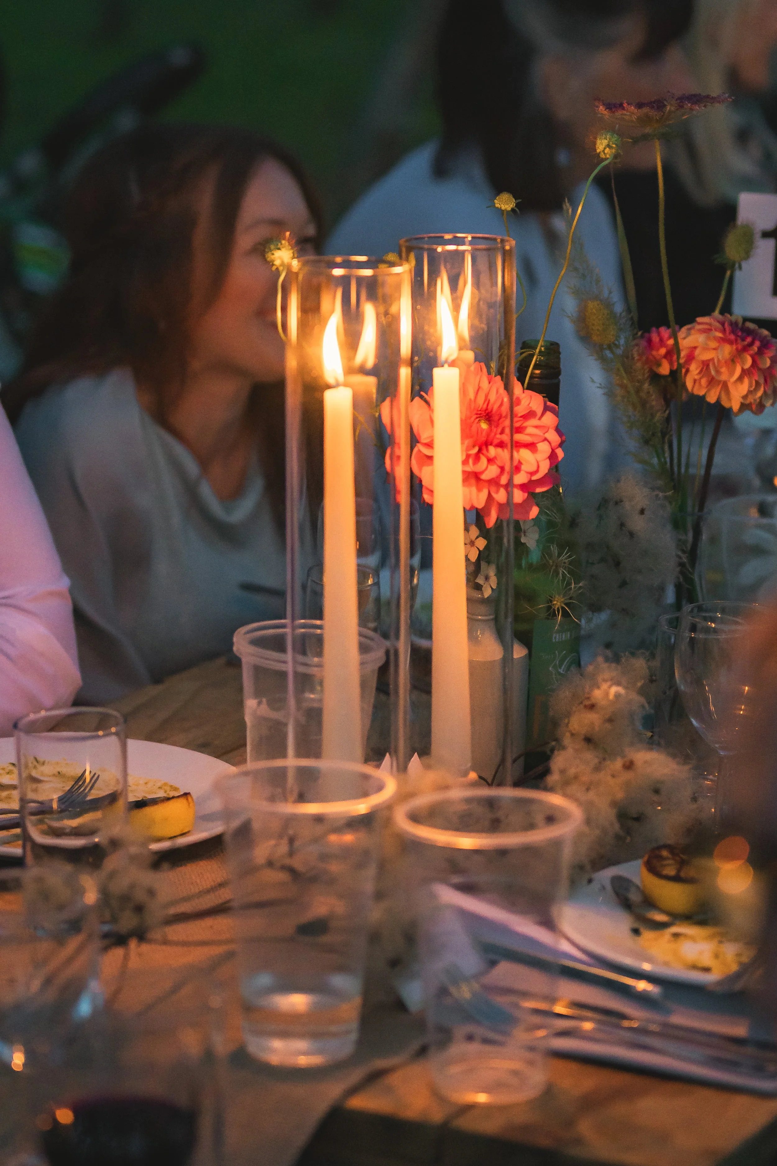 A dining table with lit candles in glass holders, flowers, plates with food, and glasses of water, set outdoors at night with people in the background.
