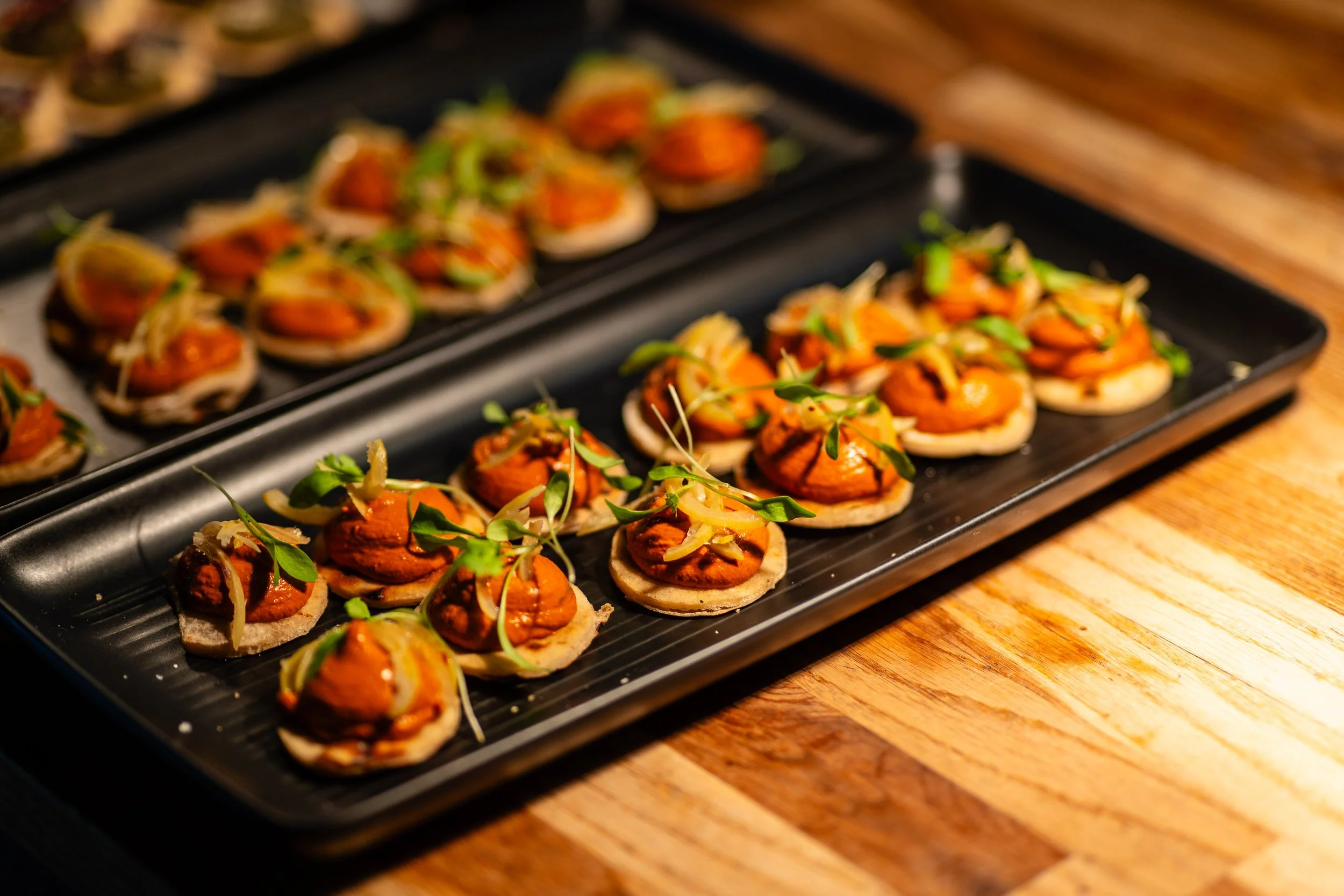 Tray of small appetizers with a base of round bread, topped with a spread, garnished with microgreens and sliced onion, on a wooden table.