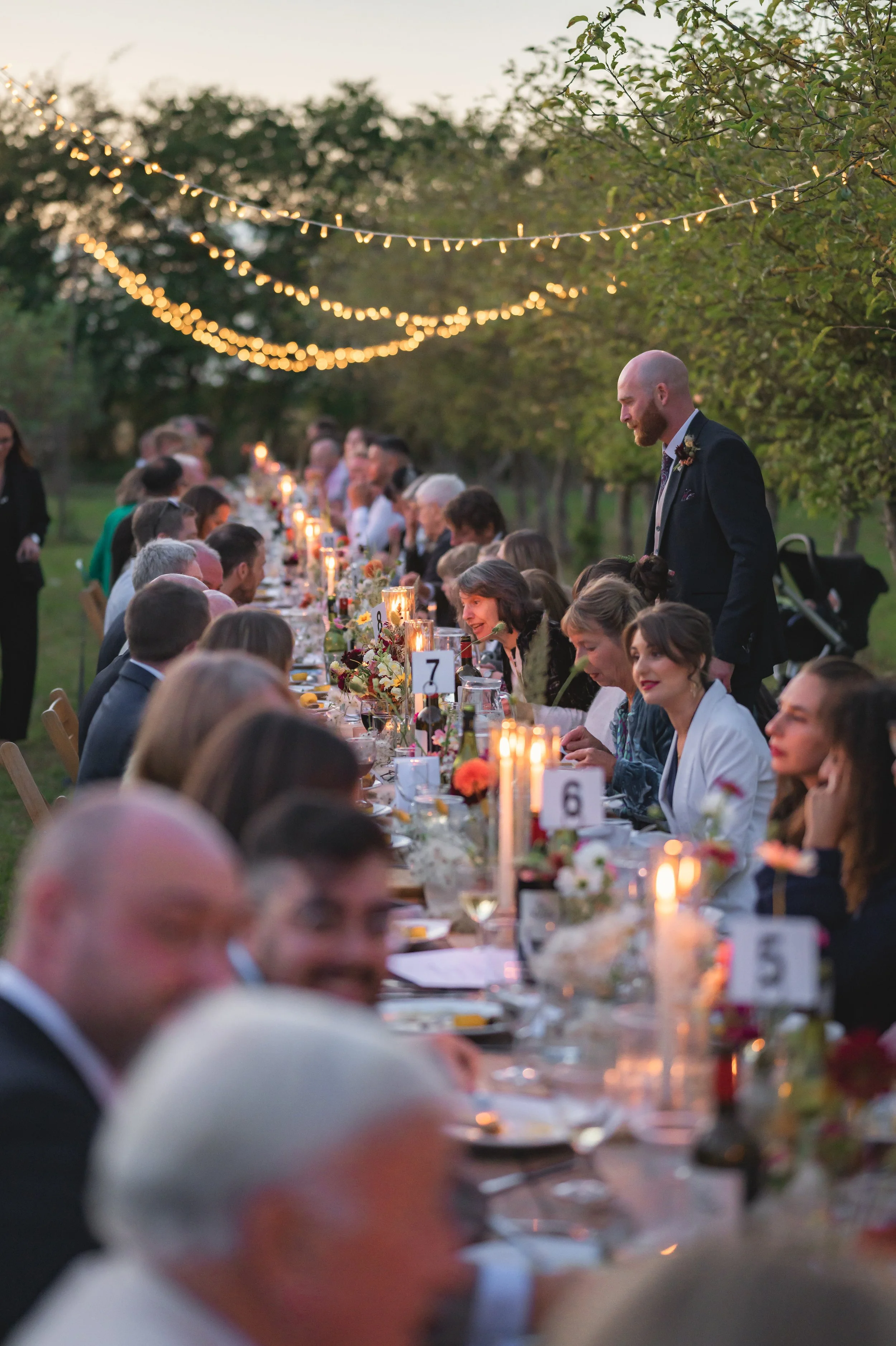 Long outdoor dinner table at dusk, decorated with candles, flowers, and string lights, with guests dressed in formal attire enjoying a celebration.