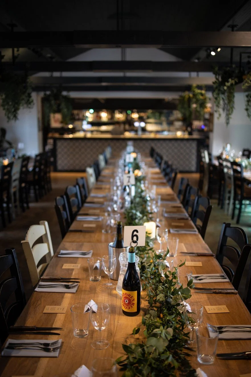 A long dining table set for a banquet with chairs on both sides, wine bottles, glassware, candlelit decorations, and green foliage as a table runner in a restaurant or event space.