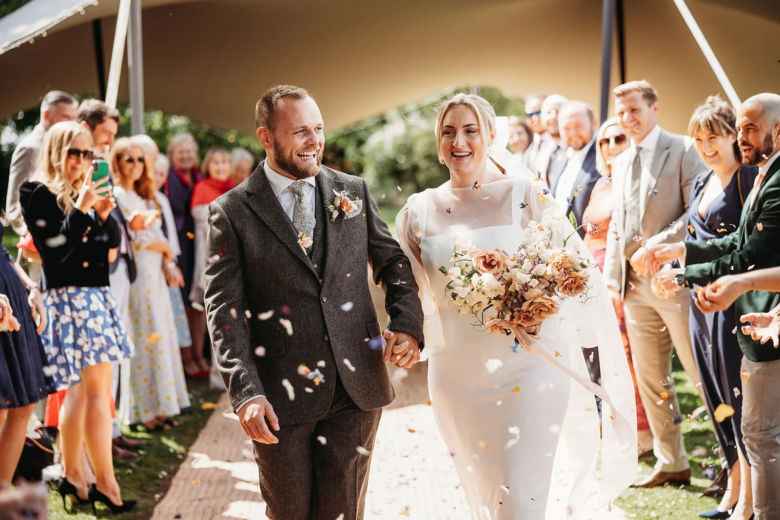 Bride and groom holding hands walking down aisle at outdoor wedding, surrounded by guests throwing confetti.