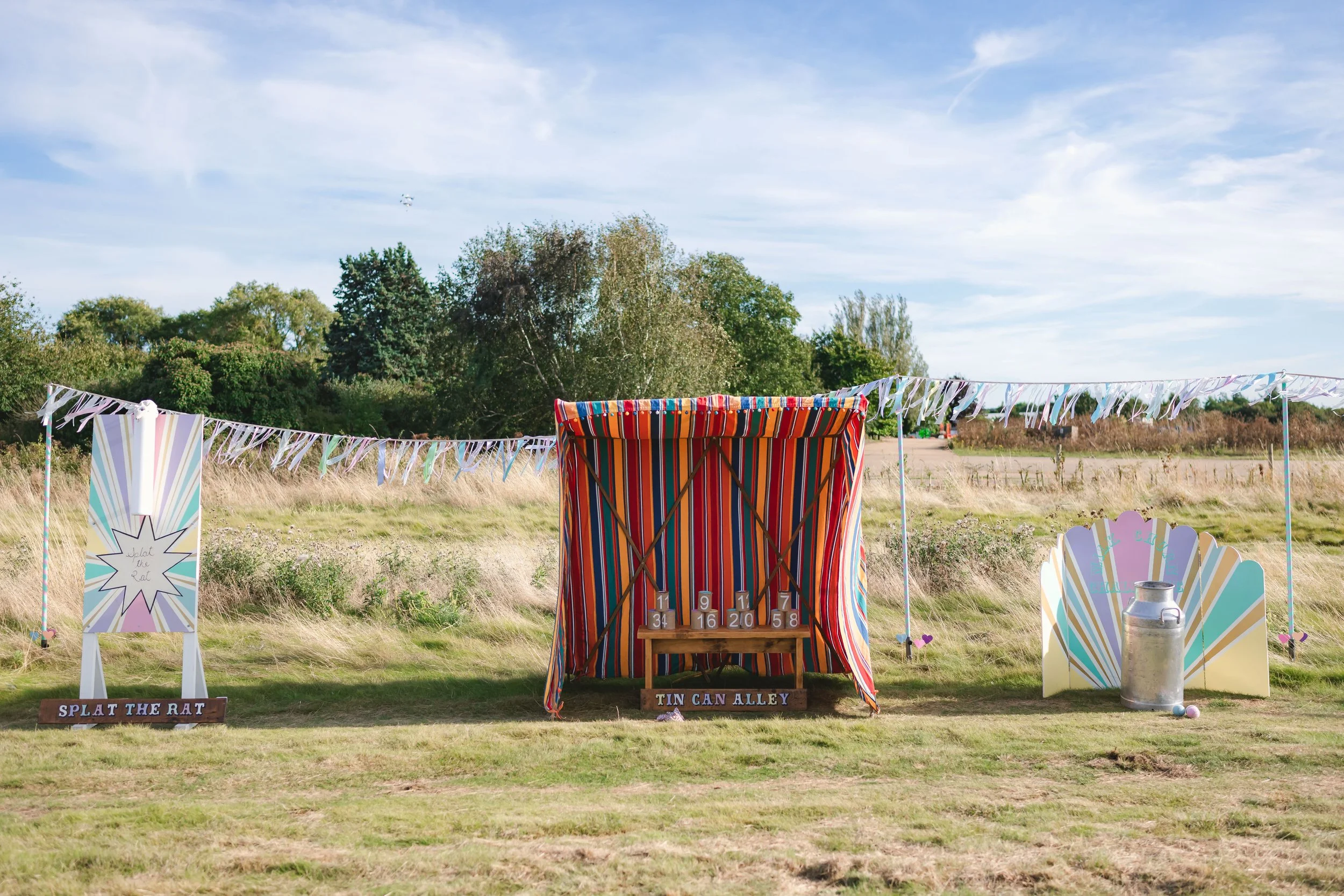 Outdoor carnival game setup with striped tent, sign that reads 'Splat the Rat', and another sign that says 'Tin Can Alley', with a milk can and painted backdrop.