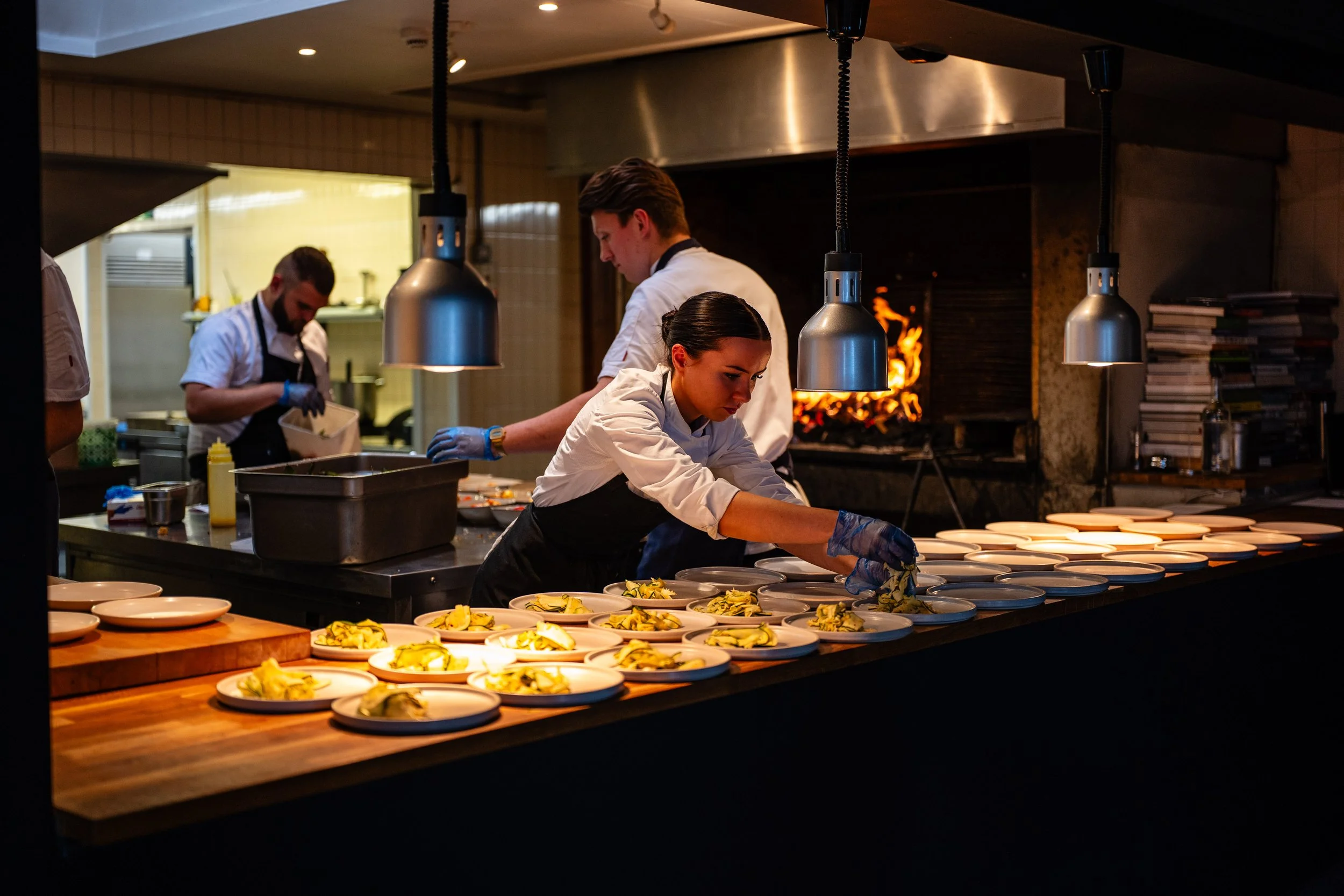 Chefs preparing and plating dishes in a professional kitchen, with a focus on a female chef arranging food on plates in front of a wood-fired oven.