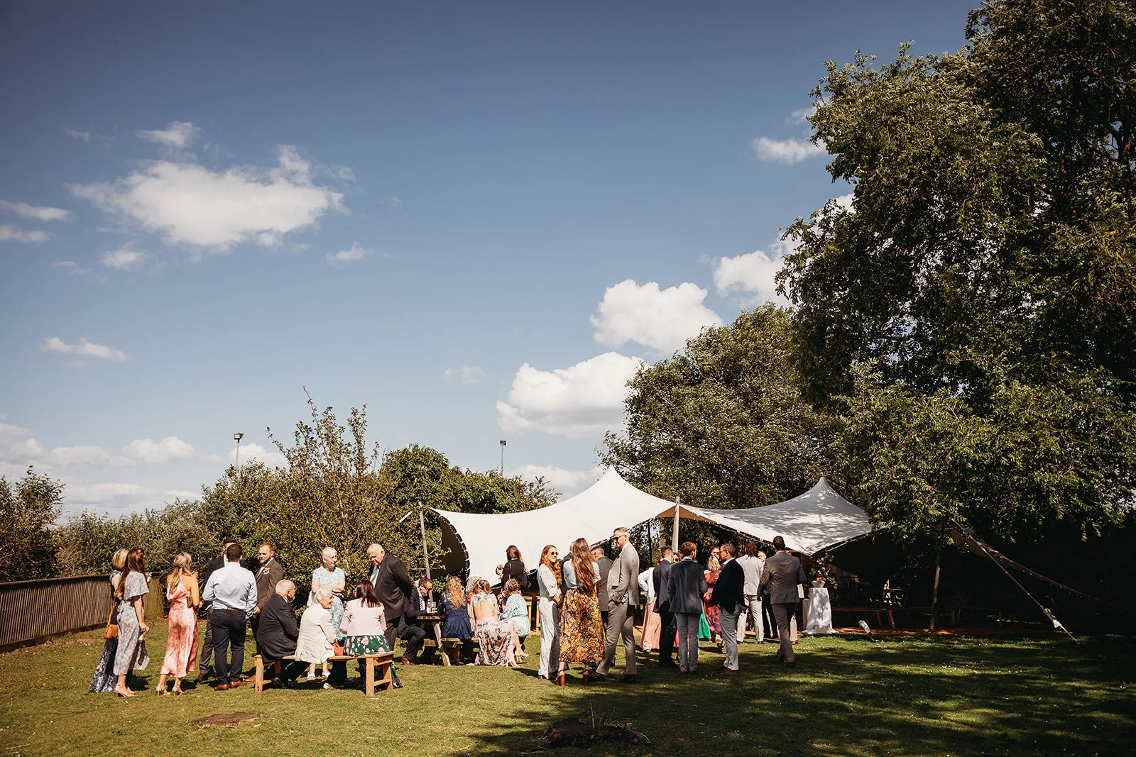 People gathered outdoors at a social event under tents, on a grassy area with trees and blue sky