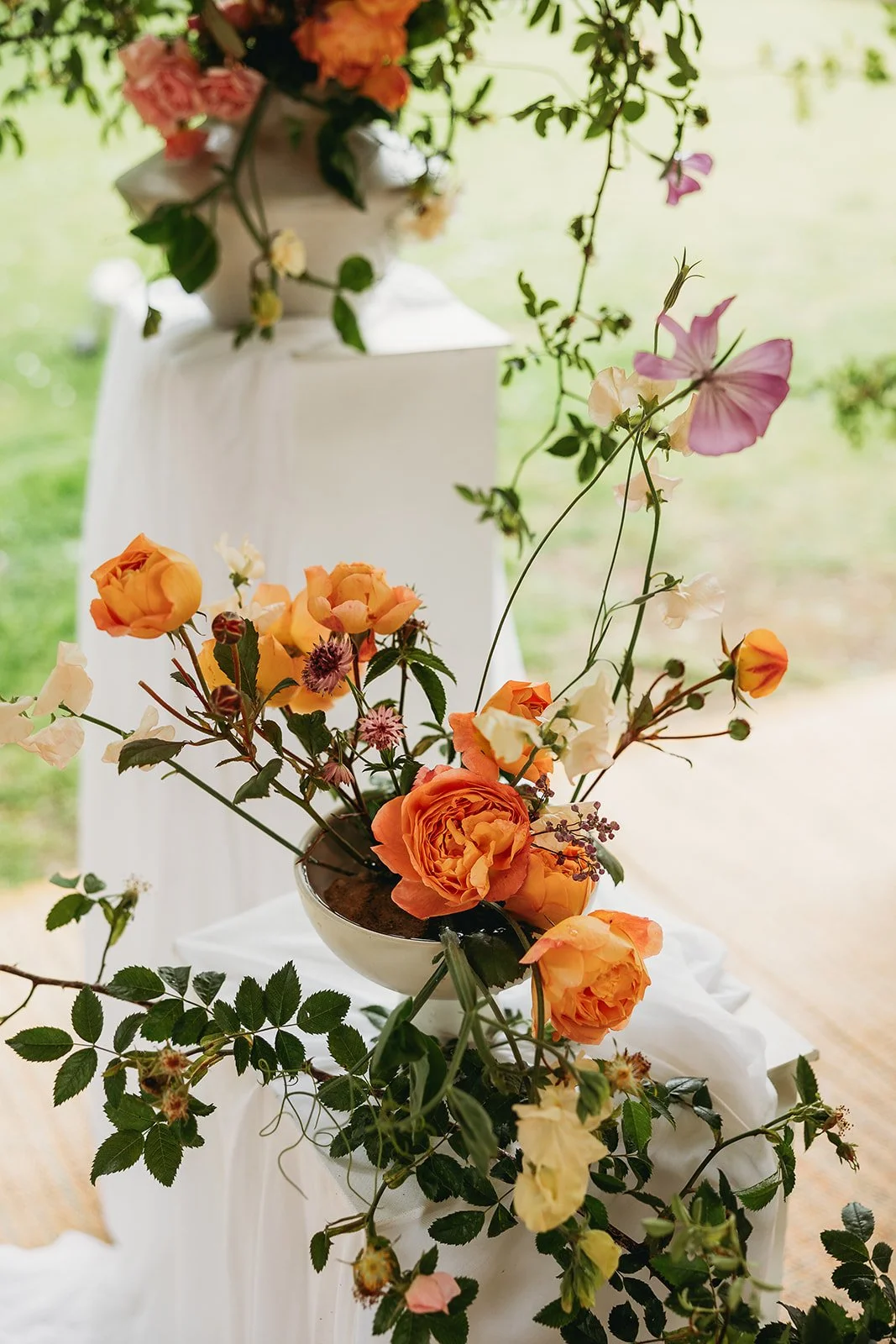 A floral arrangement with orange, pink, and yellow roses and other flowers in a white bowl on a white-covered table, with a background of greenery.