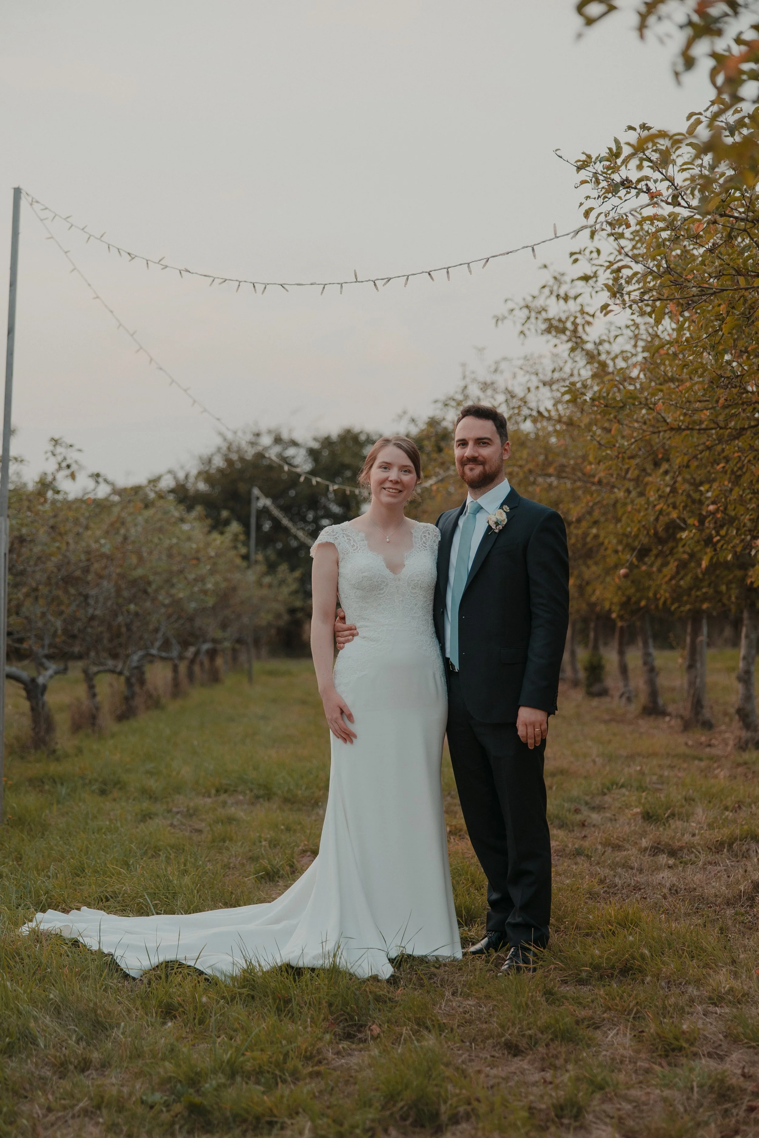 A bride and groom standing together outdoors in a vineyard, dressed in wedding attire, with string lights hanging above them and trees with autumn leaves in the background.