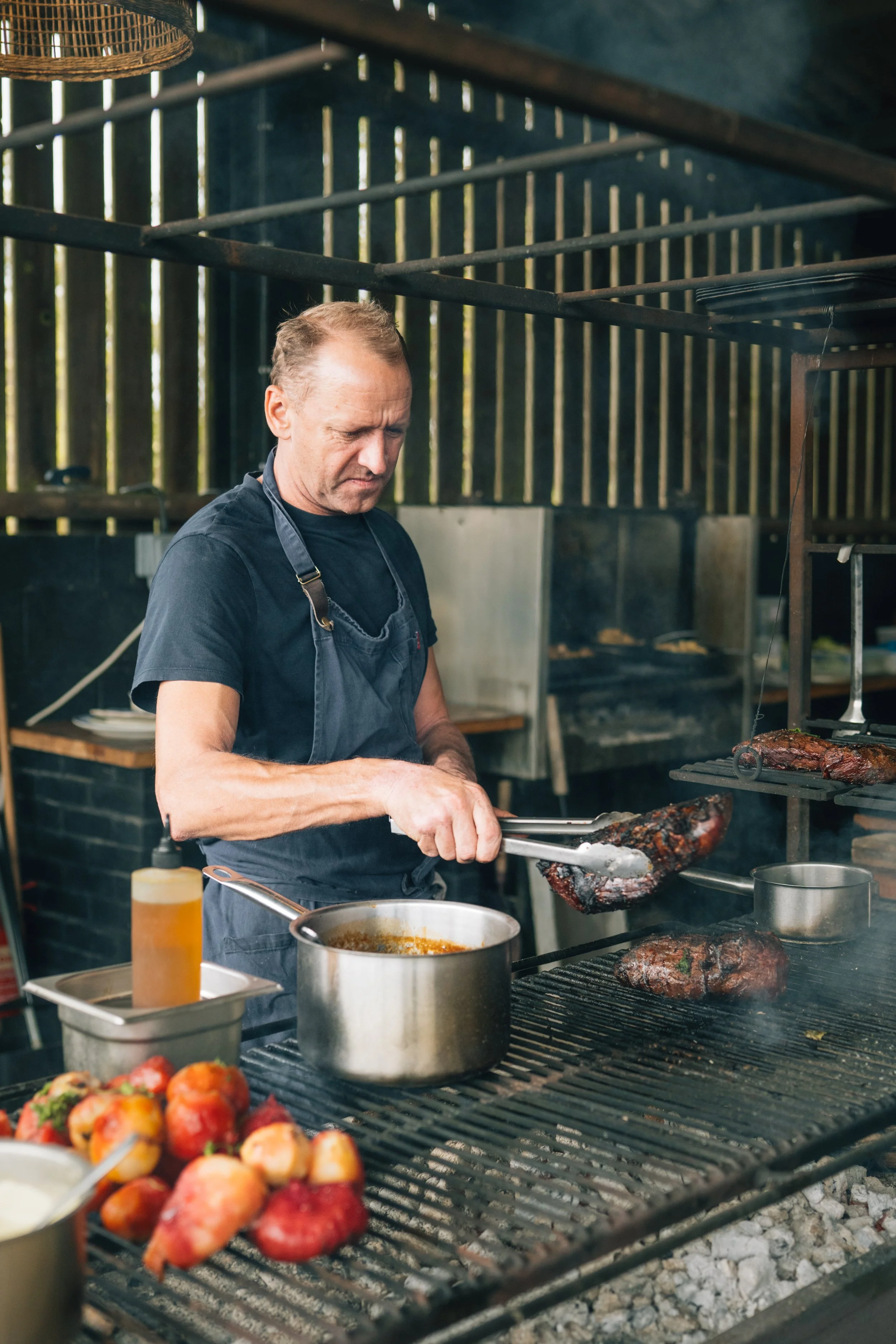 Man cooking barbecue on an outdoor grill with smoked meat, tomatoes, and sauces nearby.