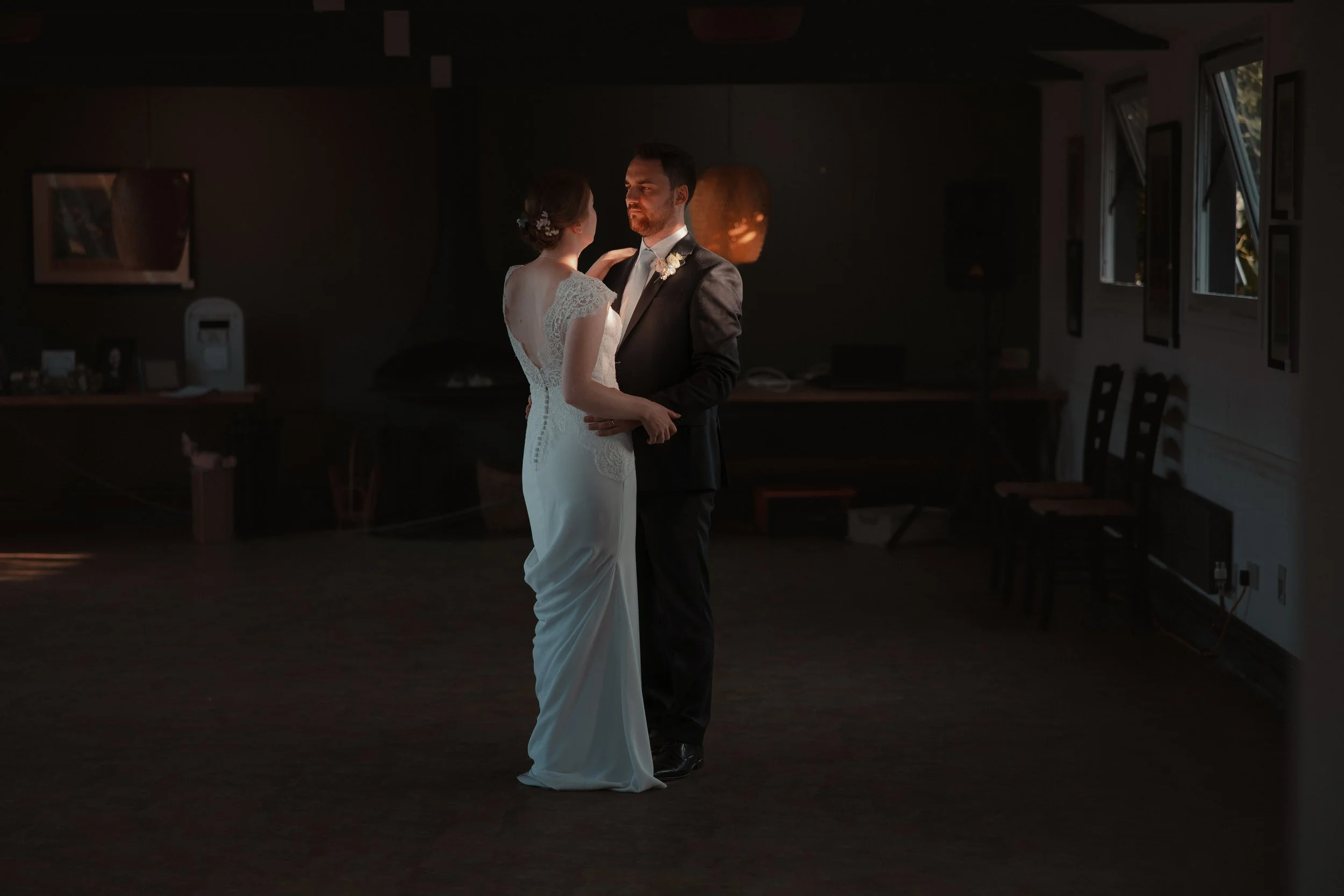 A bride and groom sharing their first dance in a dimly lit room with windows on one side, the bride in a white lace wedding dress and the groom in a black suit.