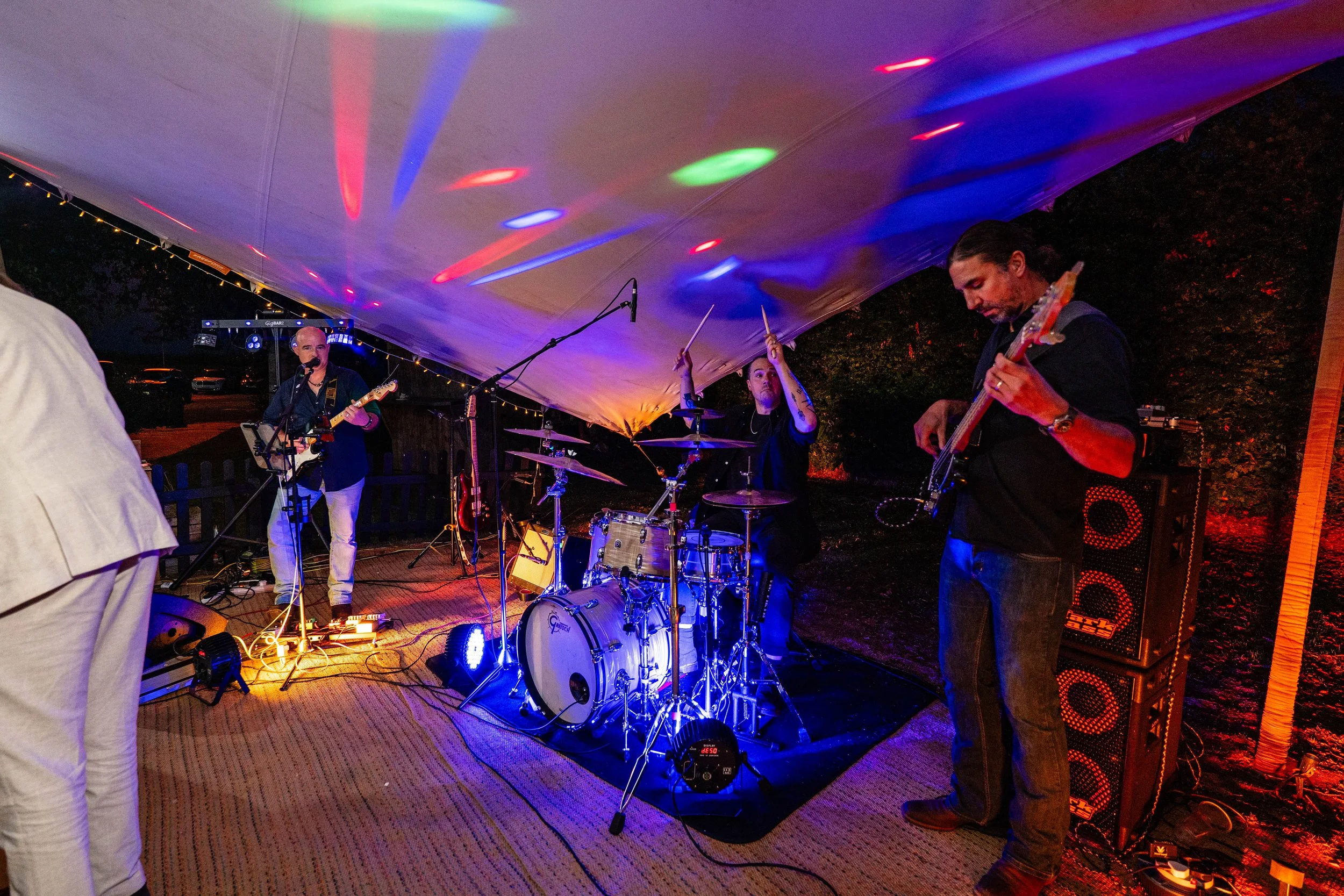 A band performing under a canopy at an outdoor nighttime event. The band includes a drummer, a guitarist, and a bassist, with colorful stage lights illuminating the scene.