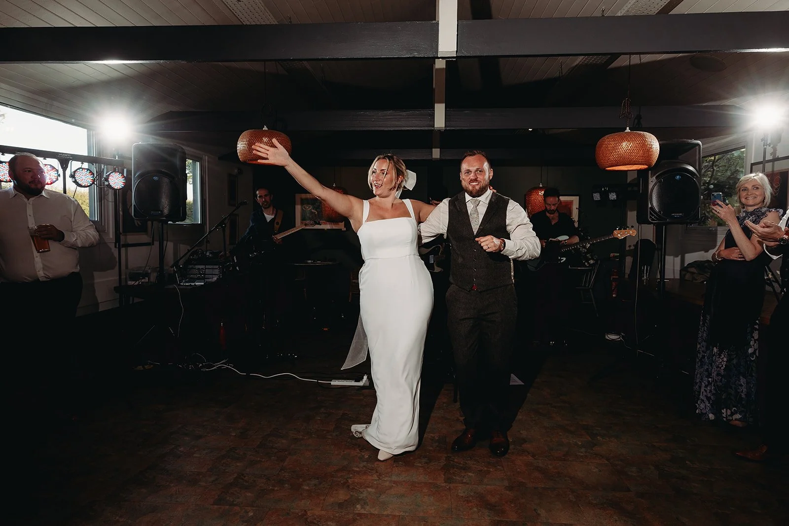 A bride and groom dancing at their wedding reception, the bride in a white gown with her arm raised, and the groom in a vest and dress shirt, smiling. There are musicians and guests in the background, some with drinks, in a decorated indoor venue wit