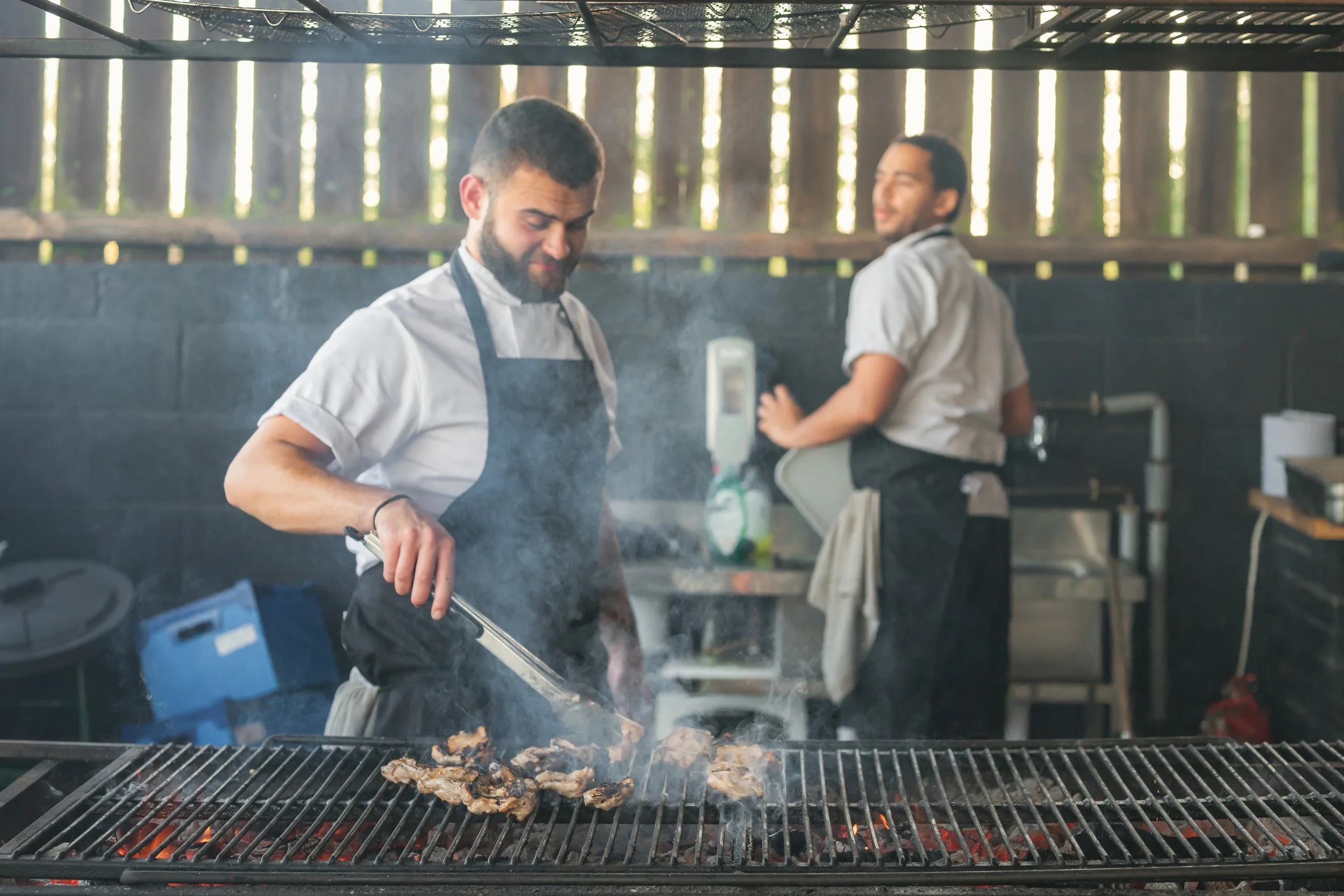 Two men grilling meat outdoors on a barbecue grill with smoke, with a wooden fence in the background.