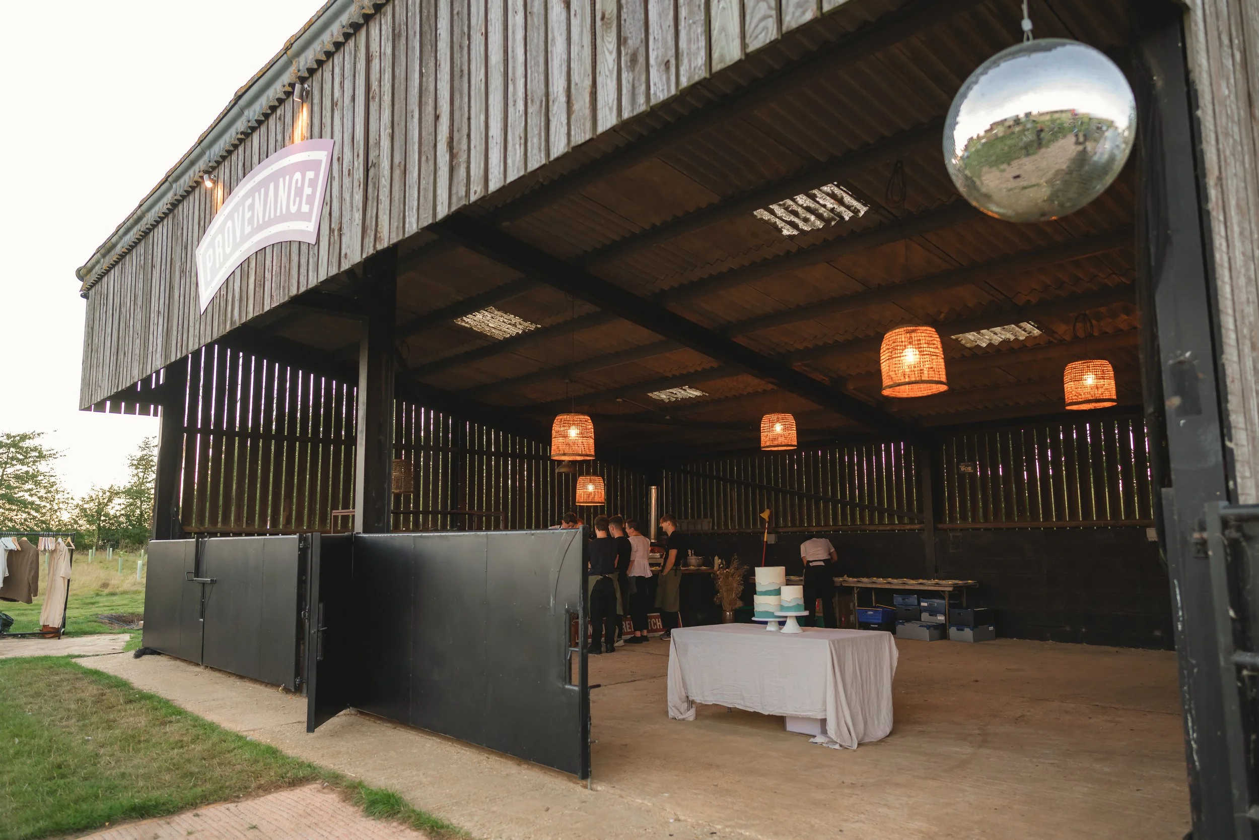 An open barn-like structure with hanging wicker lights and people gathered around a table, with a cake on a table at the front.