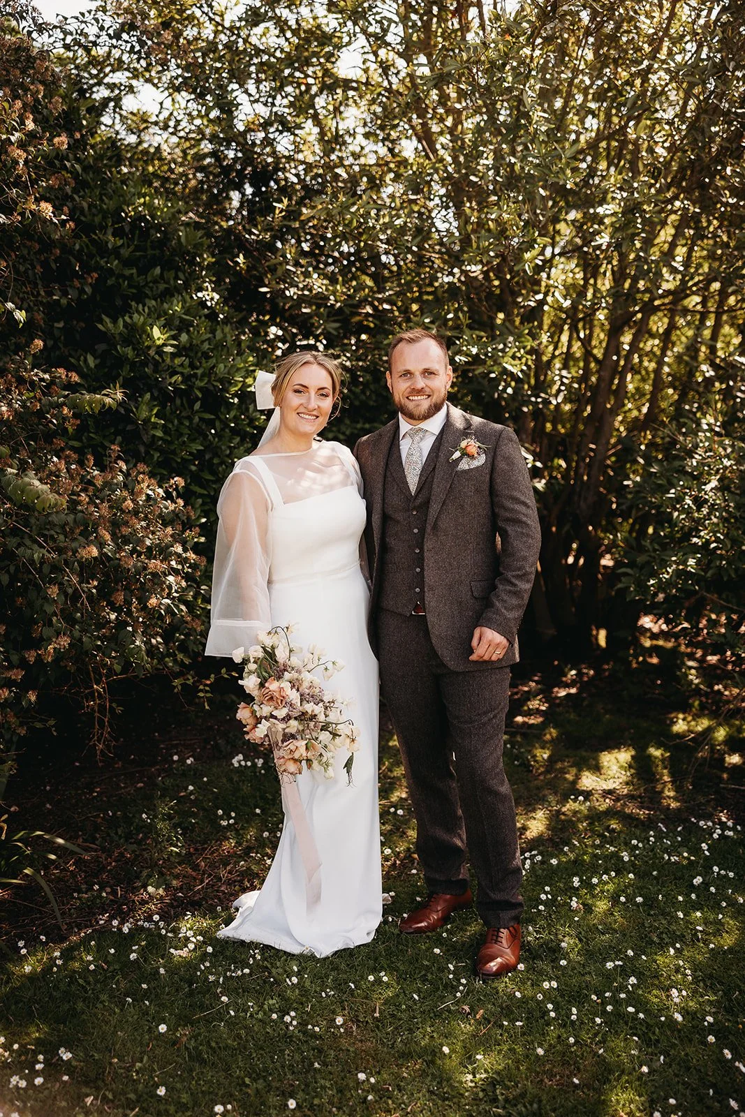 A bride and groom standing outdoors in a garden or park, smiling for a wedding photo surrounded by greenery and trees. The bride is wearing a white wedding dress with sheer long sleeves and holding a bouquet of flowers. The groom is dressed in a gray