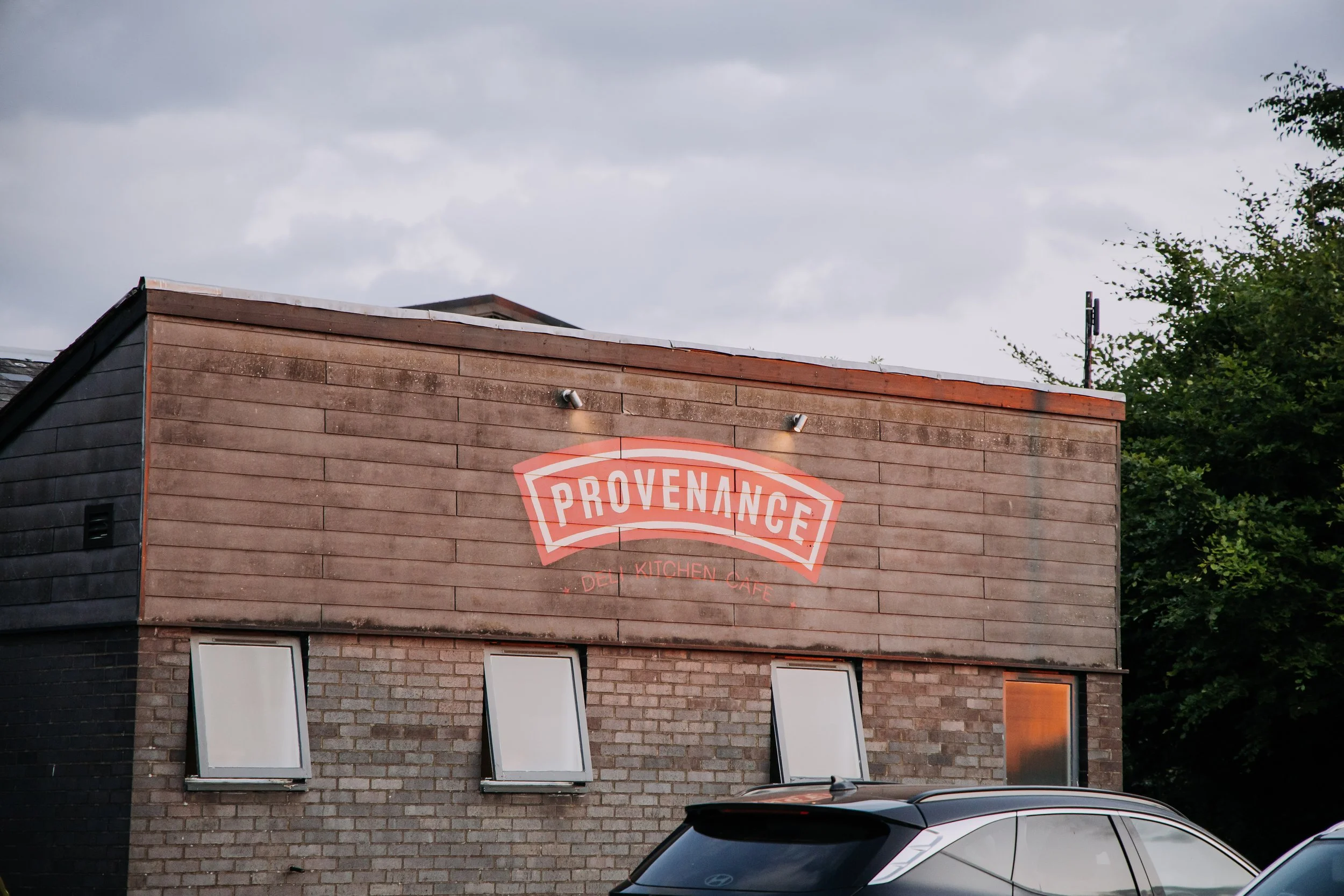 A two-story brick building with a wooden upper facade displaying a neon sign that reads "Provenance" with smaller text underneath that says "Deli Kitchen Cafe." The building has three tilted windows on the lower brick section and several lights illum