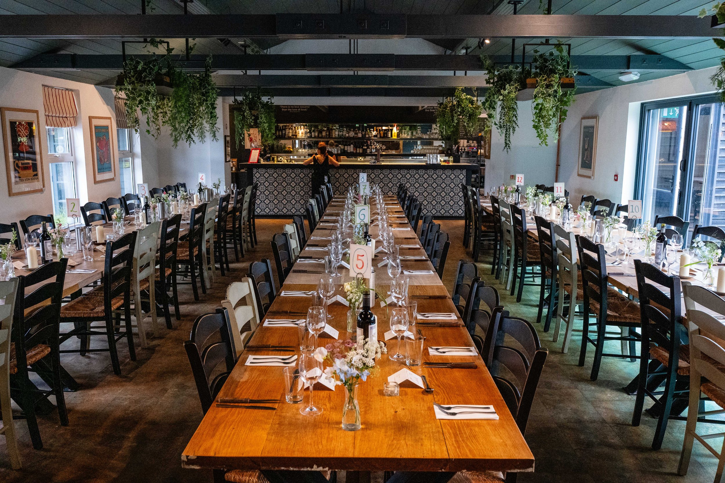 Inside a restaurant with long wooden tables set for a formal event, decorated with wine bottles, glasses, napkins, and floral centerpieces. The room has hanging plants, framed art on the walls, and a bar at the back.