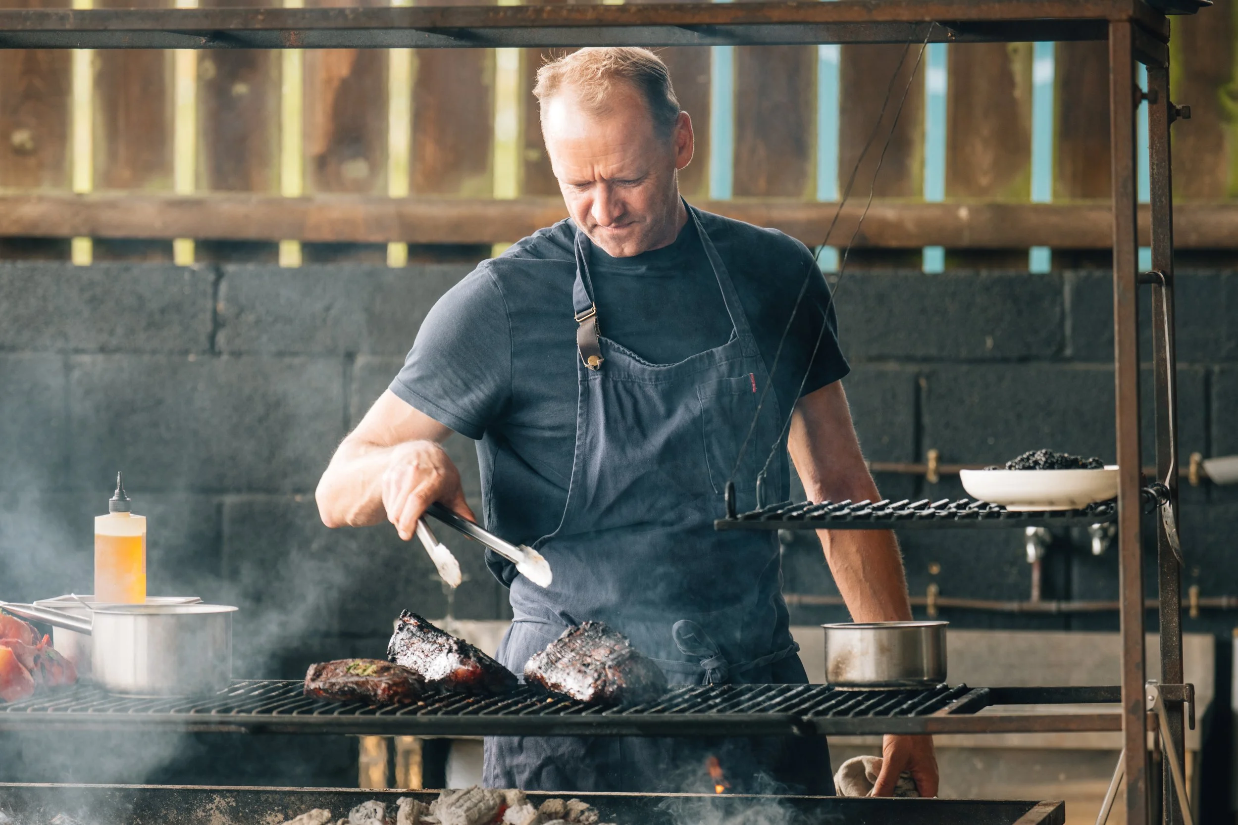 Man grilling steaks outside on a barbecue grill, using tongs, wearing a dark apron and t-shirt, with a wooden fence and trees in the background.