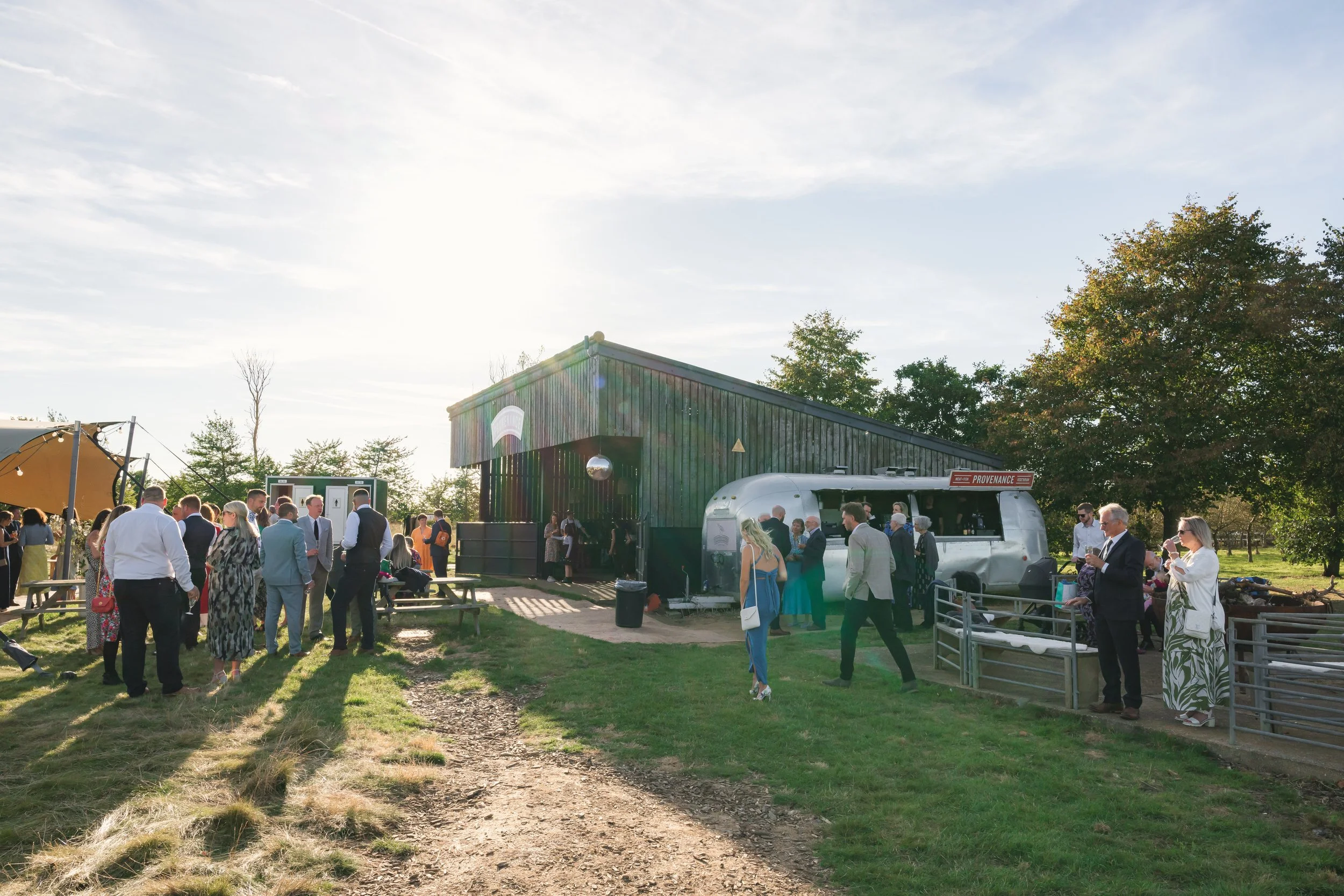 People gathering outside a rustic building with a metal trailer, some chatting and others taking photos, on a sunny day with trees in the background.